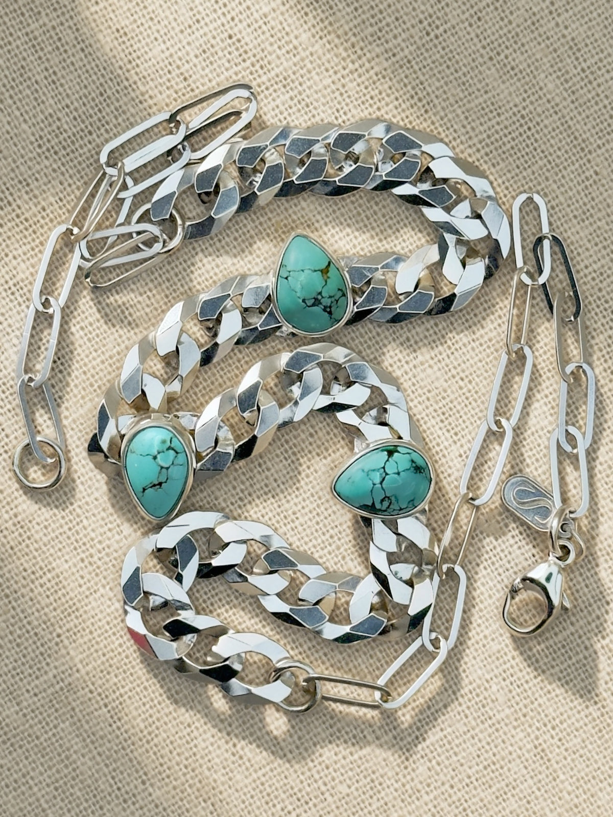 Full Blue Reign necklace coiled on cream fabric, showing curb and paperclip chains, lobster clasp, and maker’s tag.