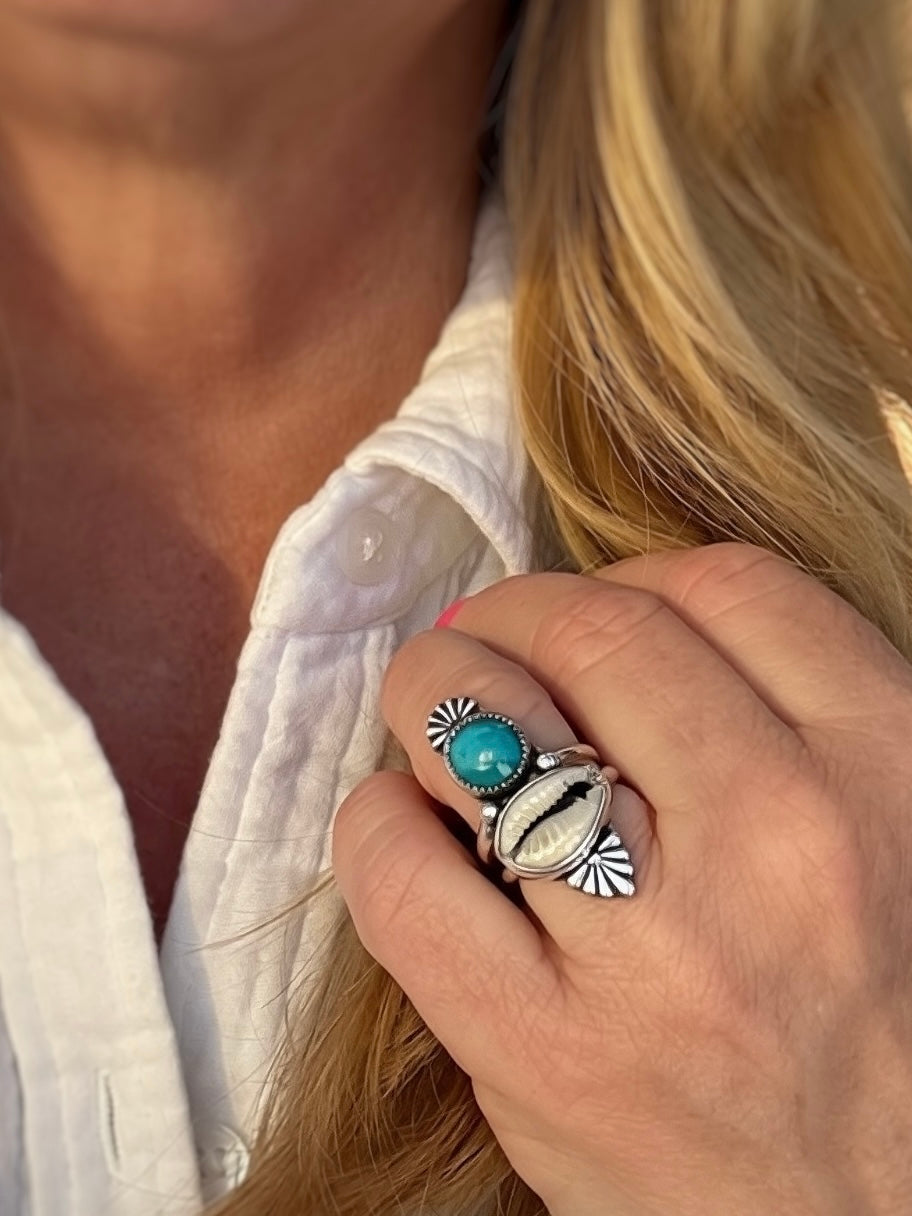 Close-up of a hand wearing a turquoise and cowrie shell ring with silver bands.