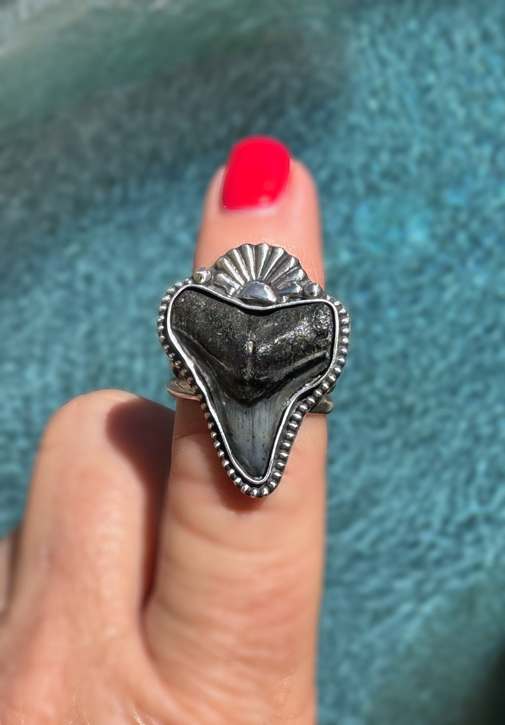 Silver ring with a shark tooth and sun design on a finger against a textured blue background
