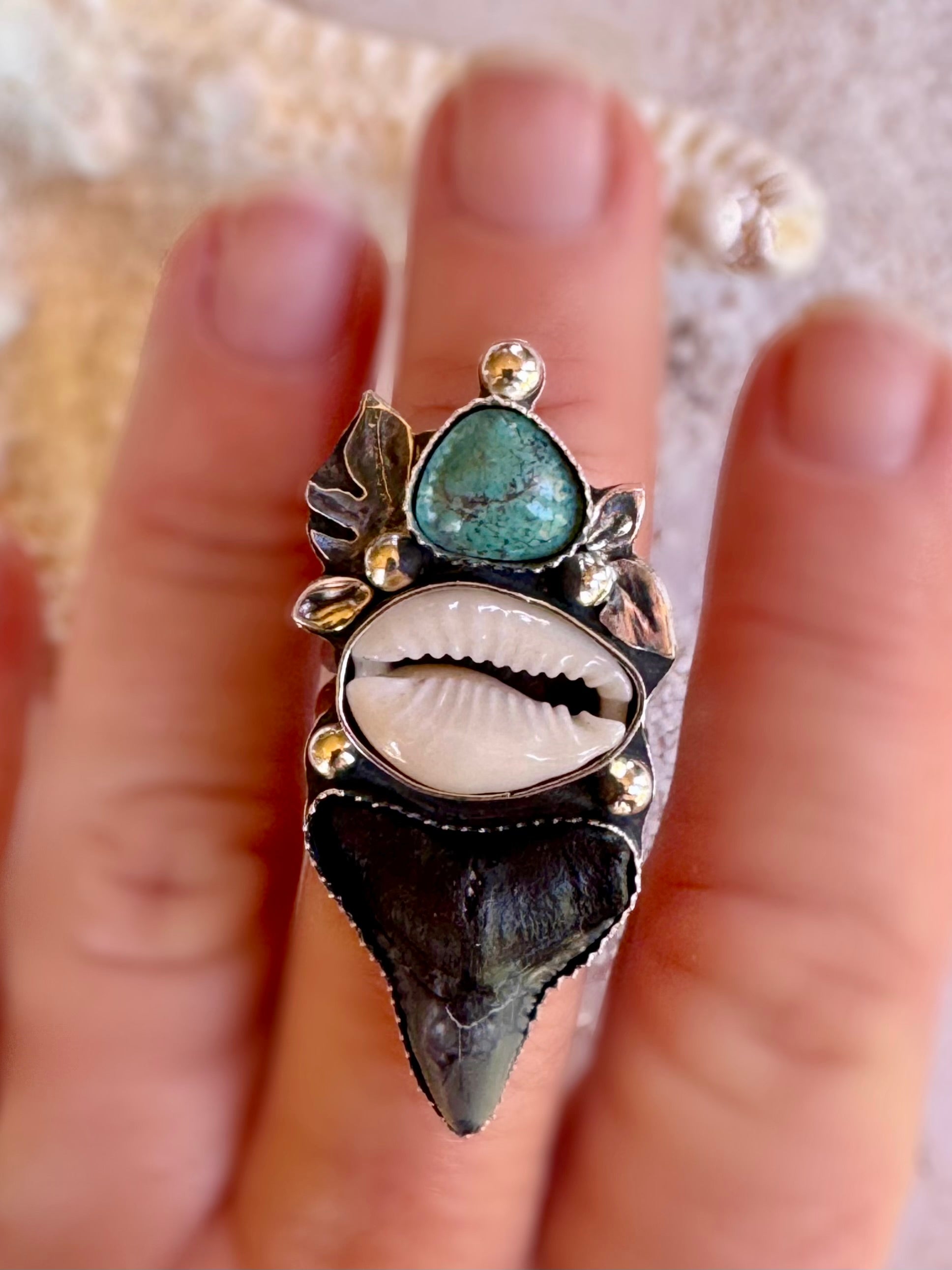 Close-up of a hand wearing an ornate ring with a shark tooth, shell and turquoise stone. Tropical leaves surround the stones.