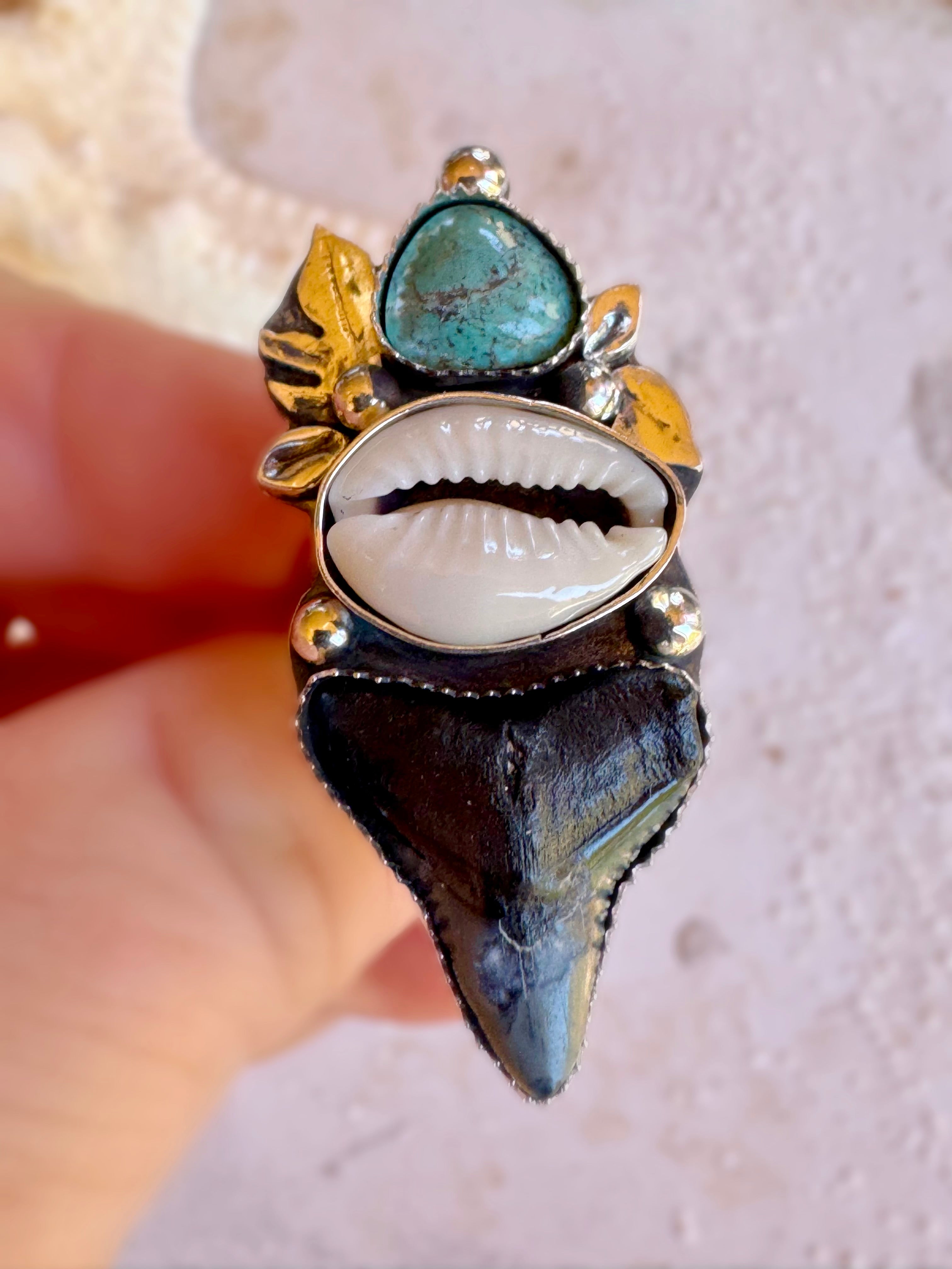 Close-up of a hand wearing a unique silver ring with shark tooth, cowrie shell and green turquoise stones and tropical leaves.