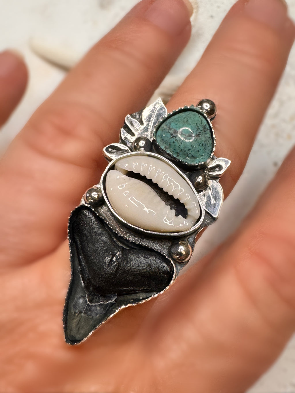 Close-up of a hand wearing a unique silver ring with shark tooth, cowrie shell and green turquoise stones.