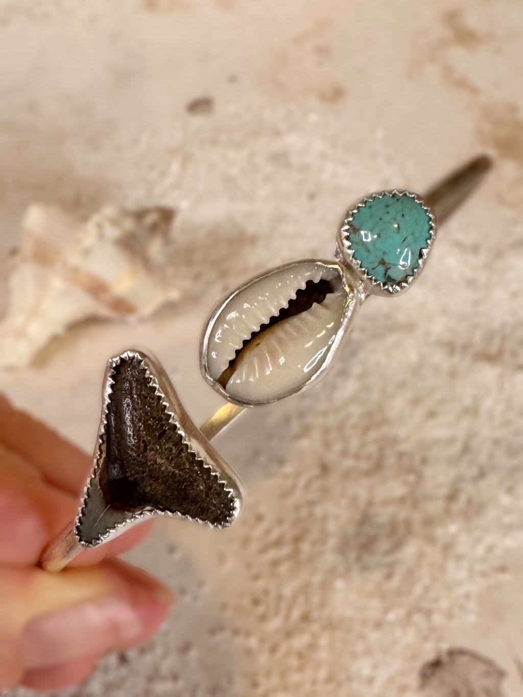 Close-up of a silver bangle with turquoise stone, shell, and shark tooth held by a hand.