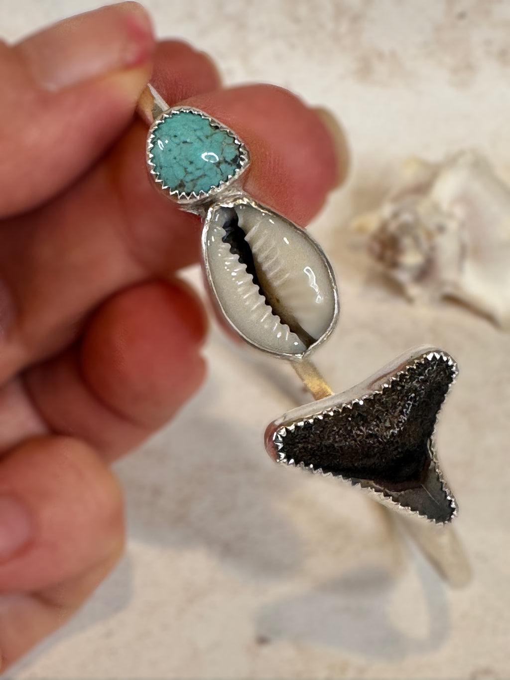 Close-up of a silver bangle with turquoise stone, shell, and shark tooth held by a hand.