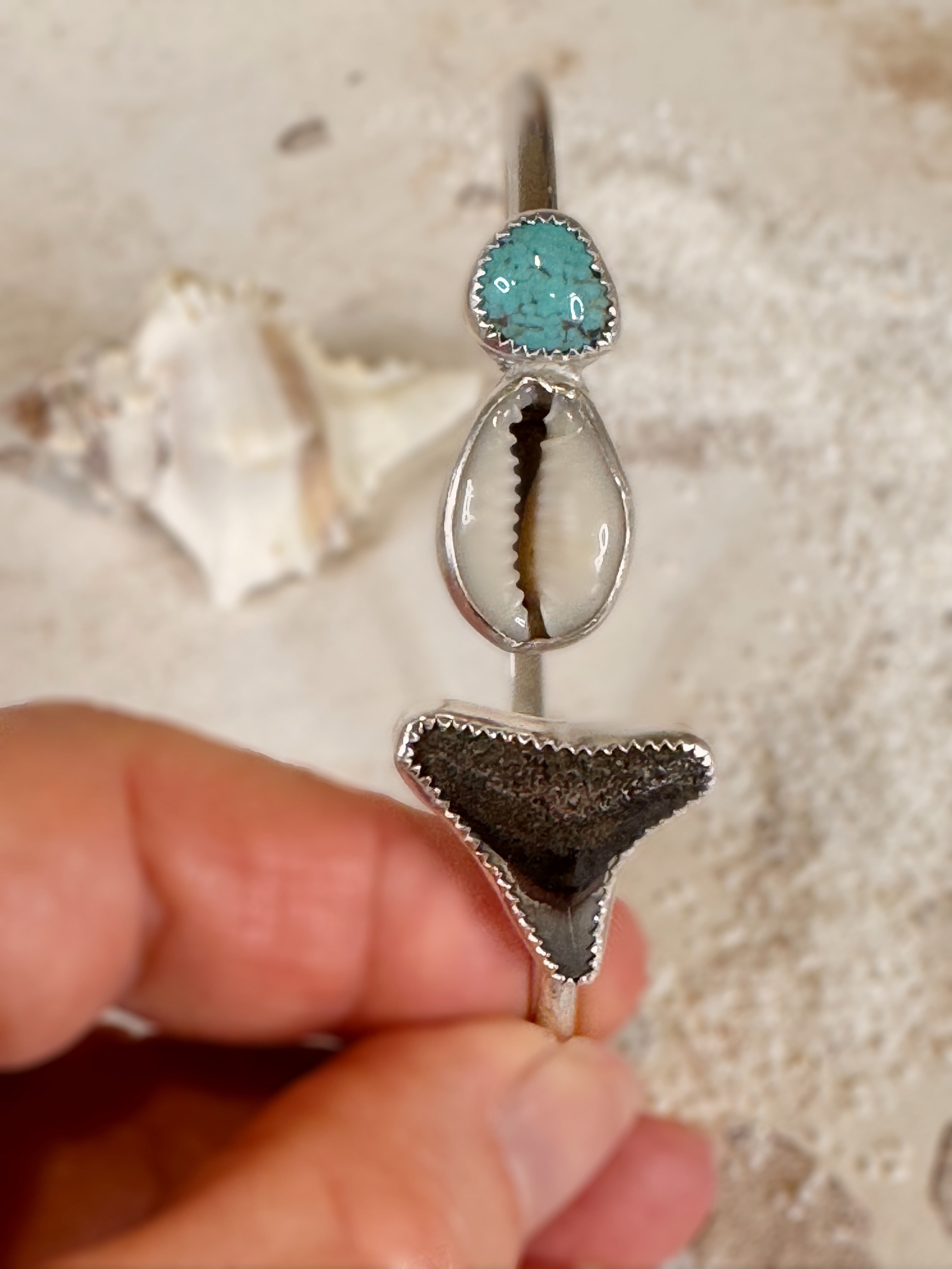 Close-up of a silver bangle with turquoise stone, shell, and shark tooth held by a hand.