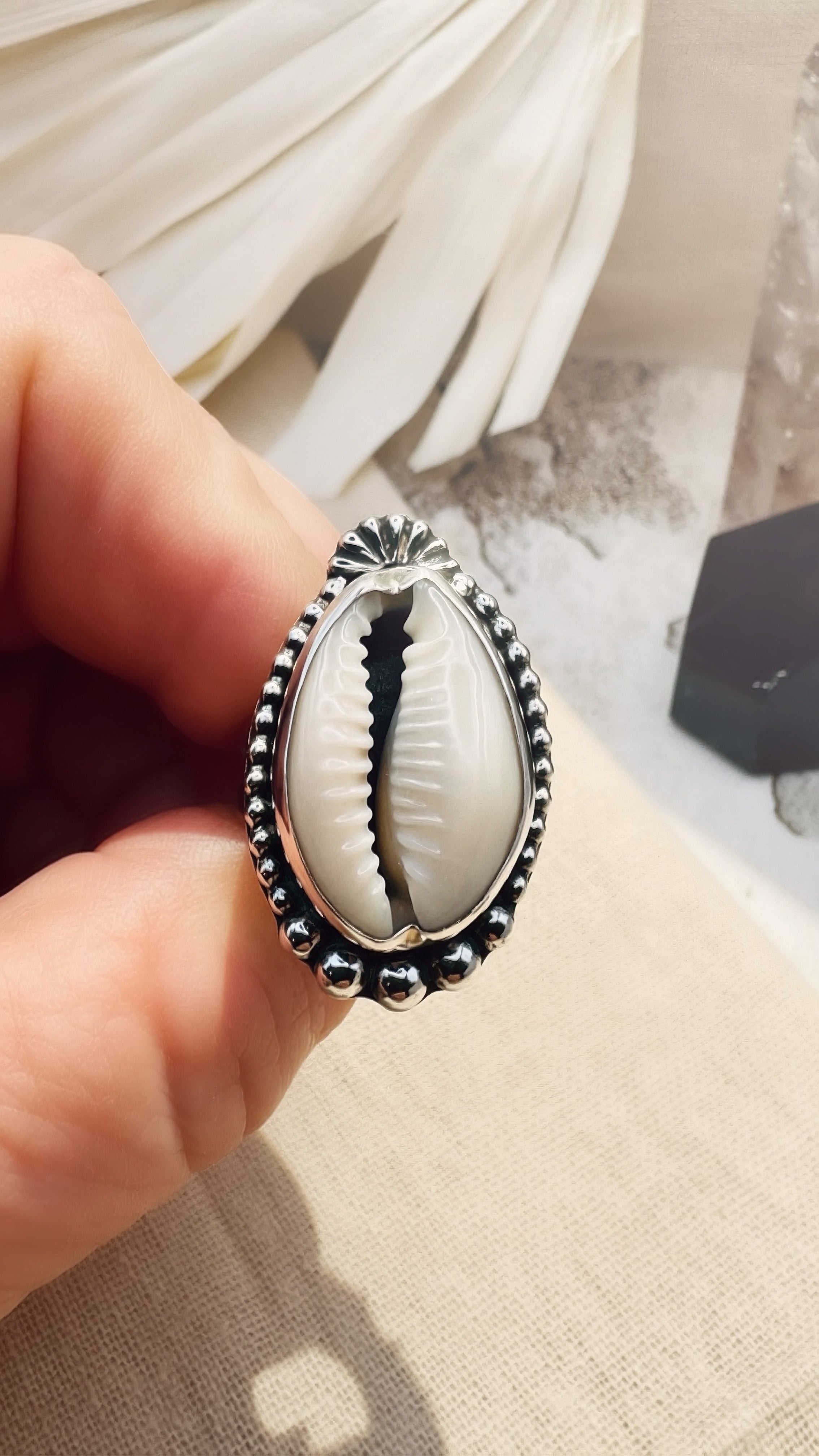 Silver ring with a cowrie shell held by a hand on a textured surface.