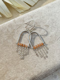 A pair of sterling silver dangle earrings with curved arches, peach moonstone beads, and dangling chain fringes, displayed on a textured beige fabric surface.
