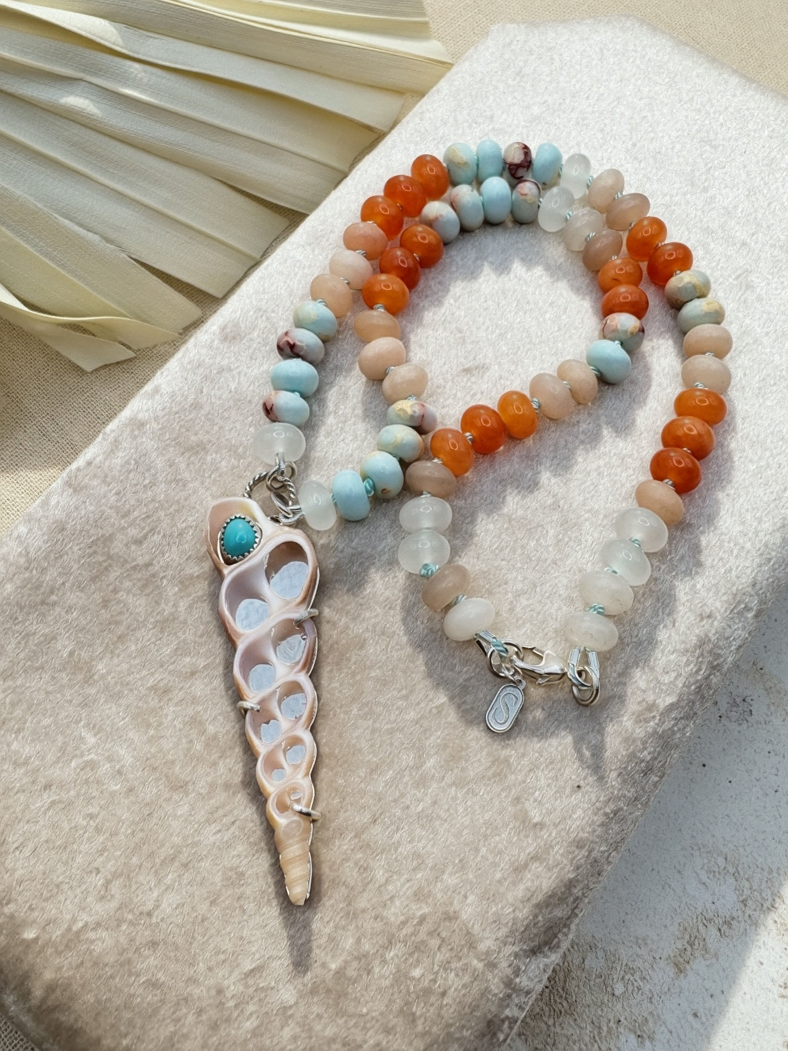 A beaded necklace with a seashell pendant and turquoise accent, displayed on a textured beige stone. The necklace features orange agate beads, light blue turquoise beads, and translucent white beads.
