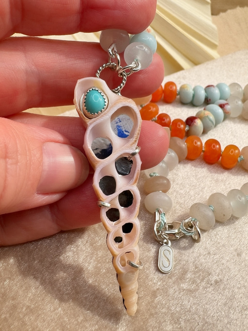 A close-up of a hand holding a seashell-pendant necklace with turquoise and blue stone accents, on a beige surface.