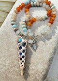 A beaded necklace with a spiral shell pendant, featuring orange beads, light blue beads, white beads, and a shell-shaped pendant with a sleeping beauty bezel set stone, displayed on a textured beige surface.