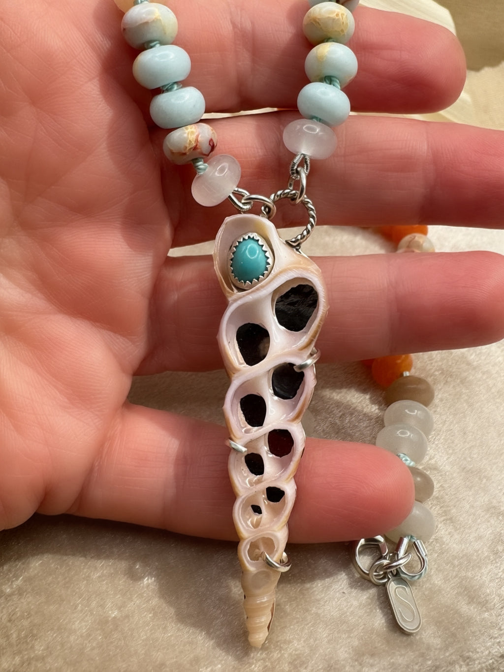 A handmade beaded necklace with a central shell pendant, held in a hand. The necklace features light blue and white beads, a silver chain, and a central shell charm and a turquoise stone.