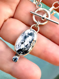 Close-up photo of sterling silver necklace with oval White Buffalo stone and teal Montana sapphire accent on a 16-inch toggle chain.