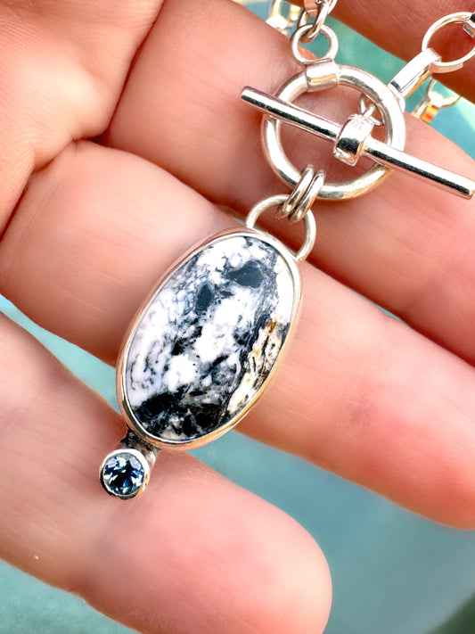 Close-up photo of sterling silver necklace with oval White Buffalo stone and teal Montana sapphire accent on a 16-inch toggle chain.