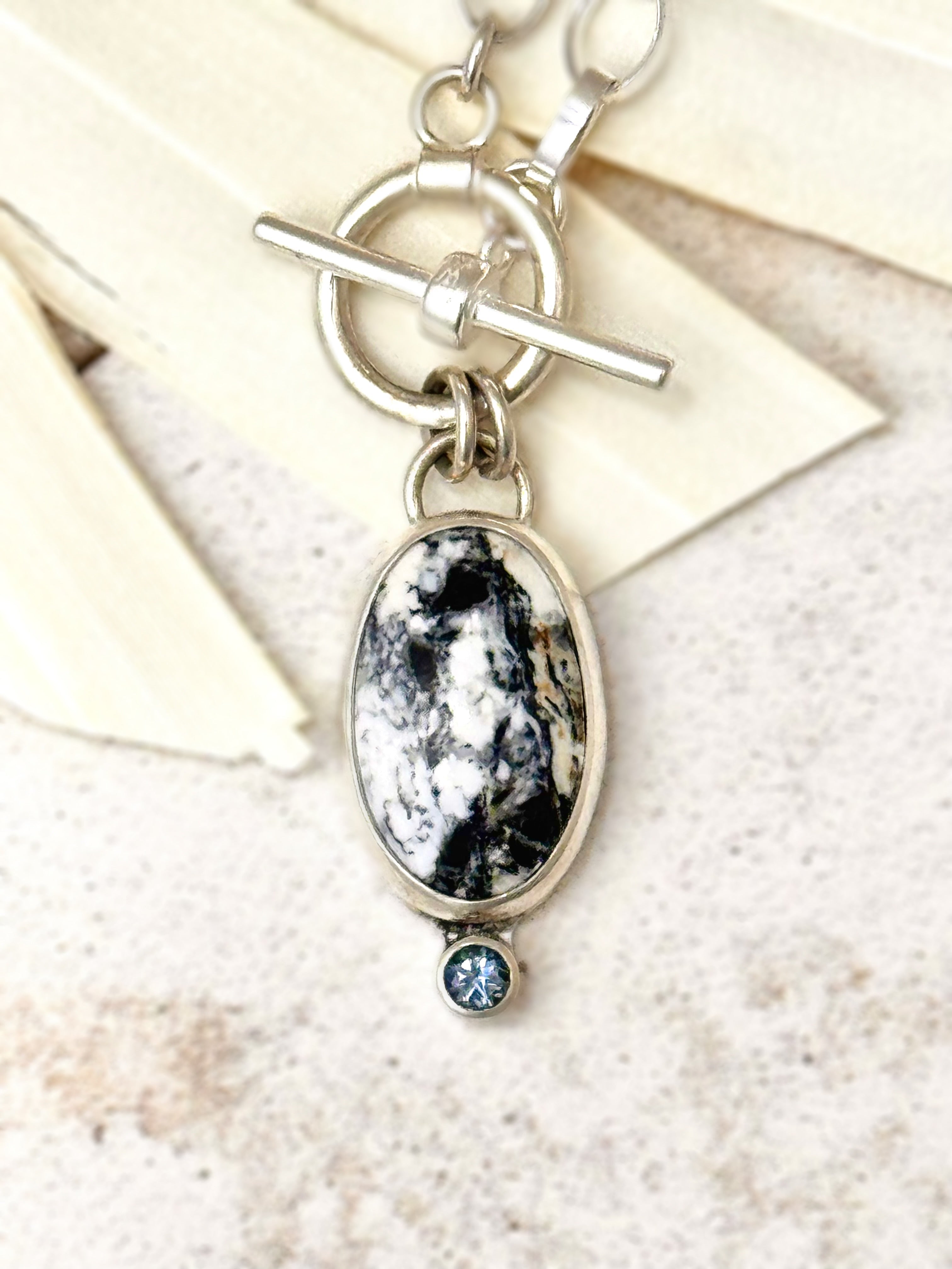 Close-up photo of sterling silver necklace with oval White Buffalo stone and teal Montana sapphire accent on a 16-inch toggle chain.