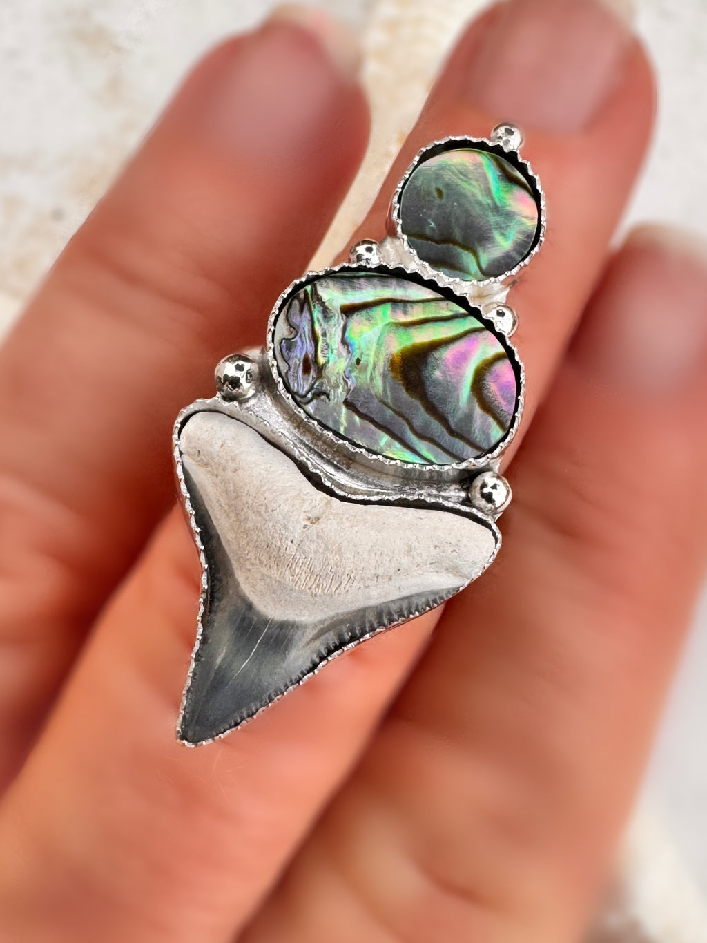 Silver ring with abalone shell and a shark tooth on a finger against a natural background