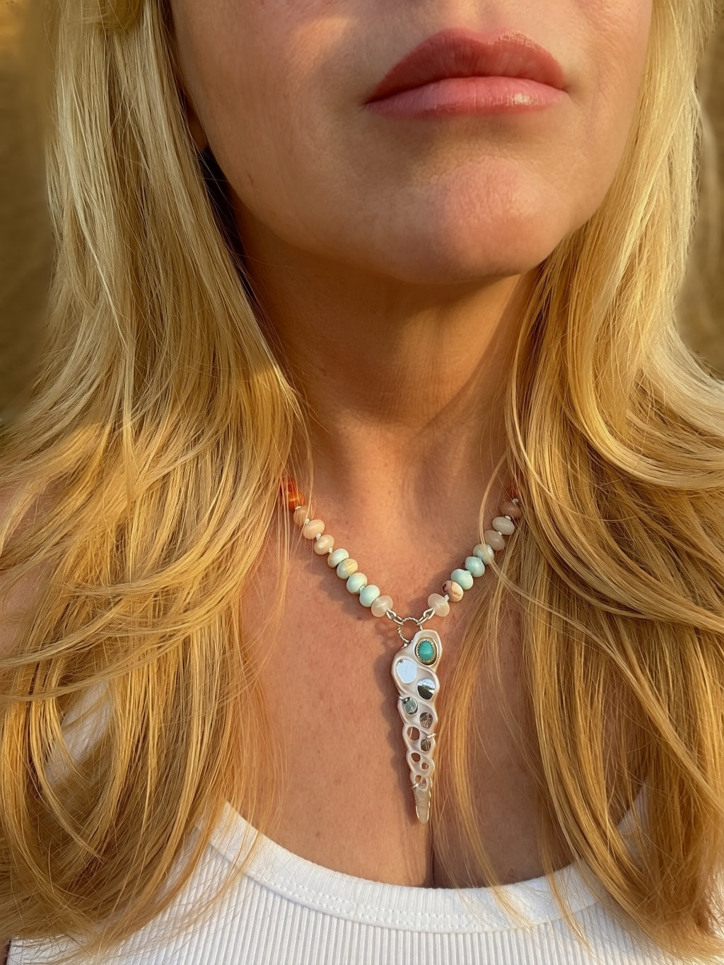 A close-up of a woman wearing a necklace with knotted pastel beads and a shell and sleeping beauty turquoise pendant, framed by blonde hair.