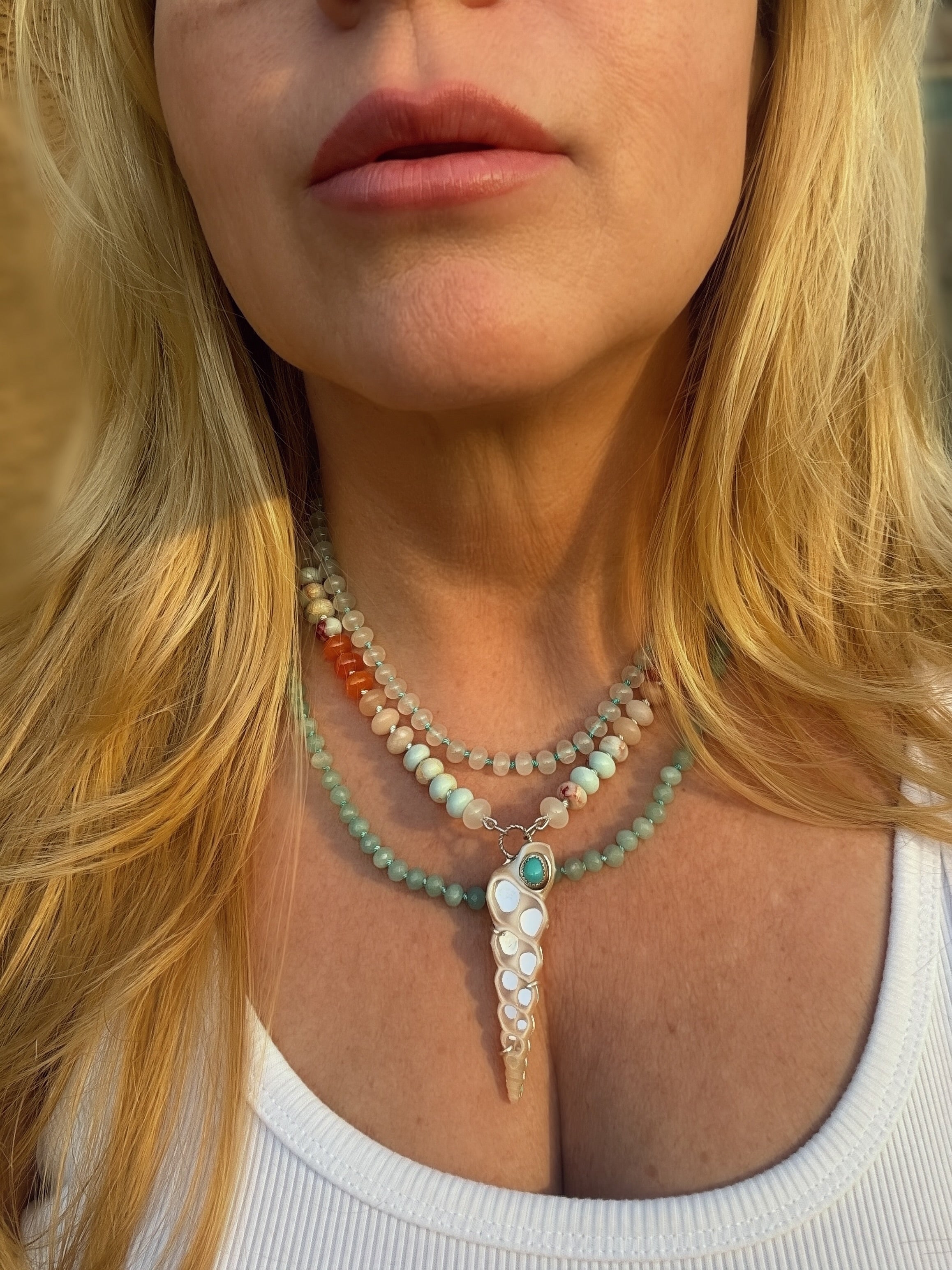 A close-up of a woman wearing a necklace withheld knotted pastel beads and a shell and sleeping beauty turquoise pendant, and 2 beaded layered necklaces, featuring seafoam and translucent white beads framed by blonde hair.