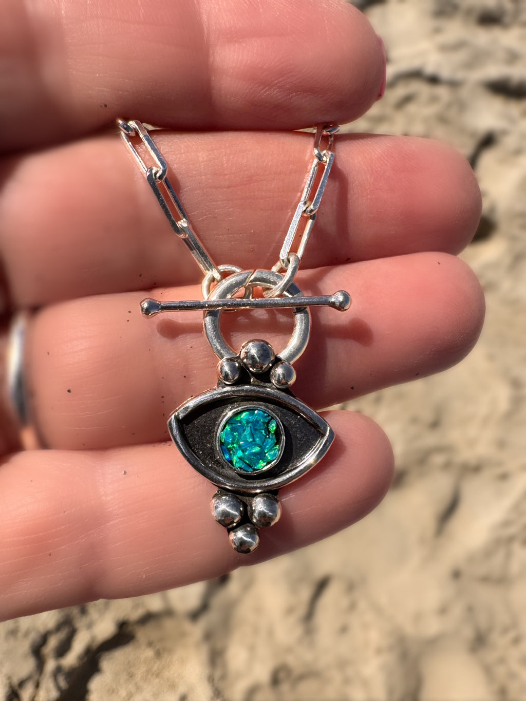 Silver necklace with an opal inlay in blue and green pendant held in a hand against a sandy background