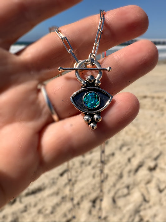 Silver pendant with blue gemstone held in a hand on a beach.