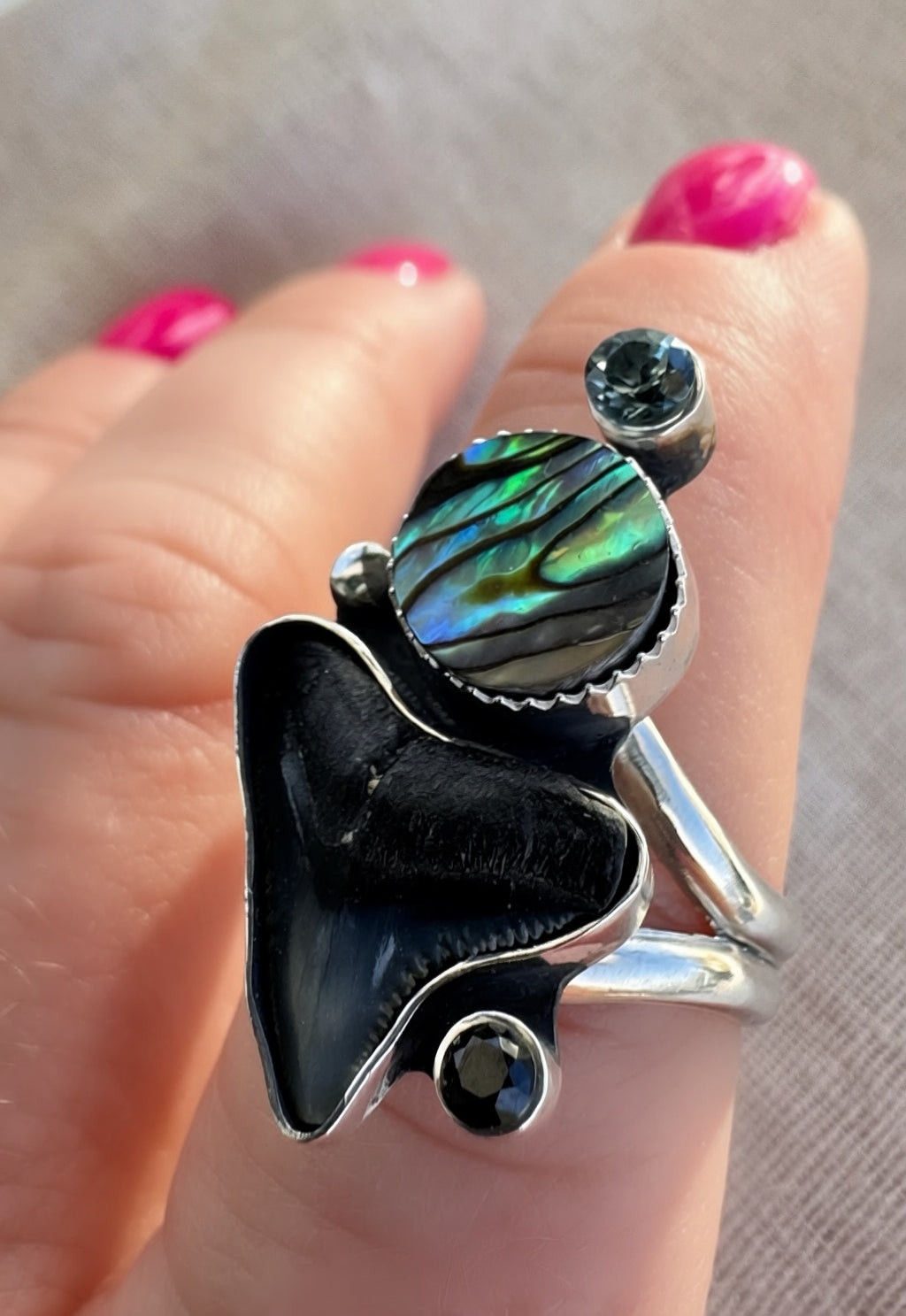 Close-up of faceted spinel gemstone above iridescent abalone shell and fossilized shark tooth set in oxidized sterling silver with polished highlights