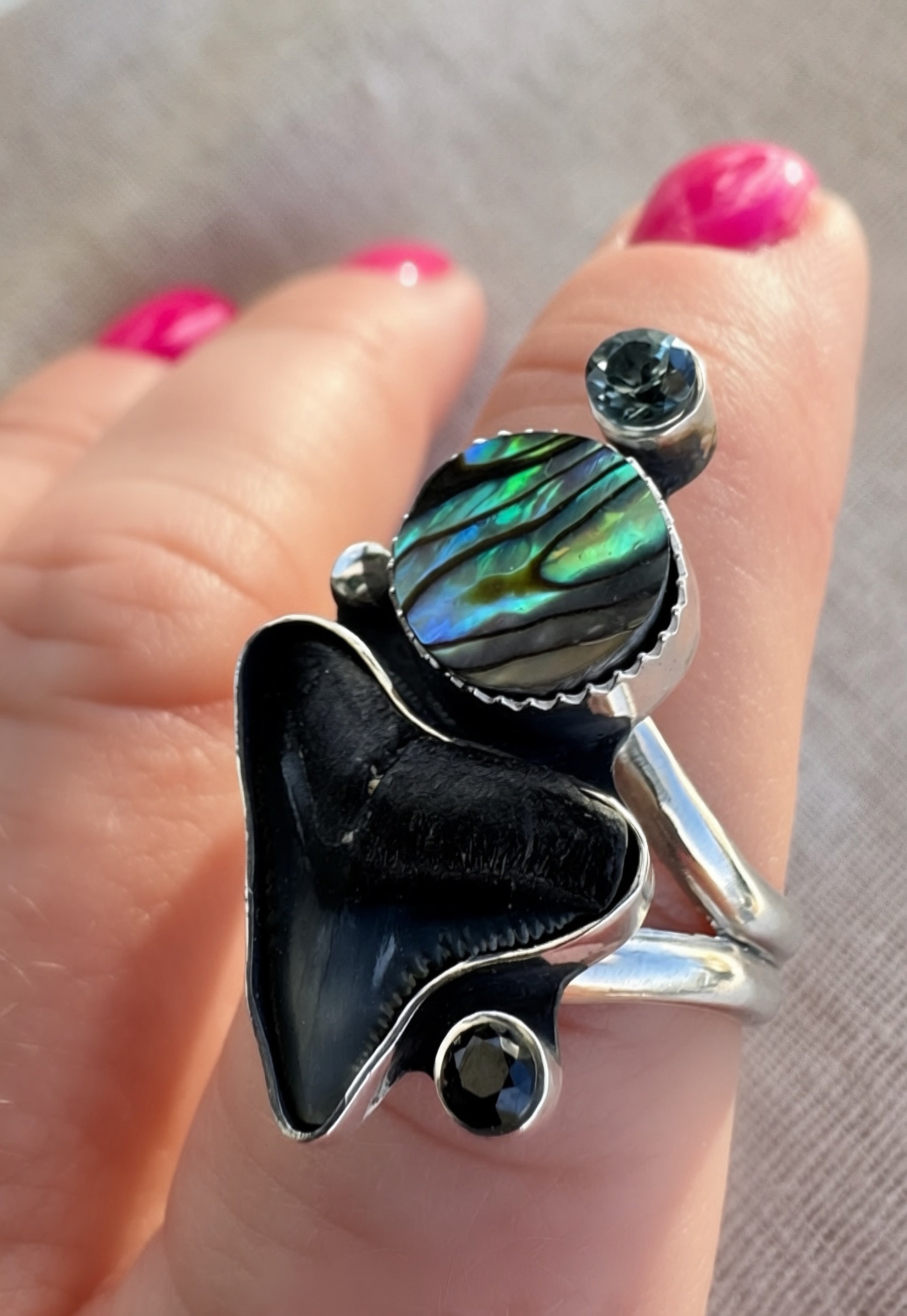 Close-up of faceted spinel gemstone above iridescent abalone shell and fossilized shark tooth set in oxidized sterling silver with polished highlights