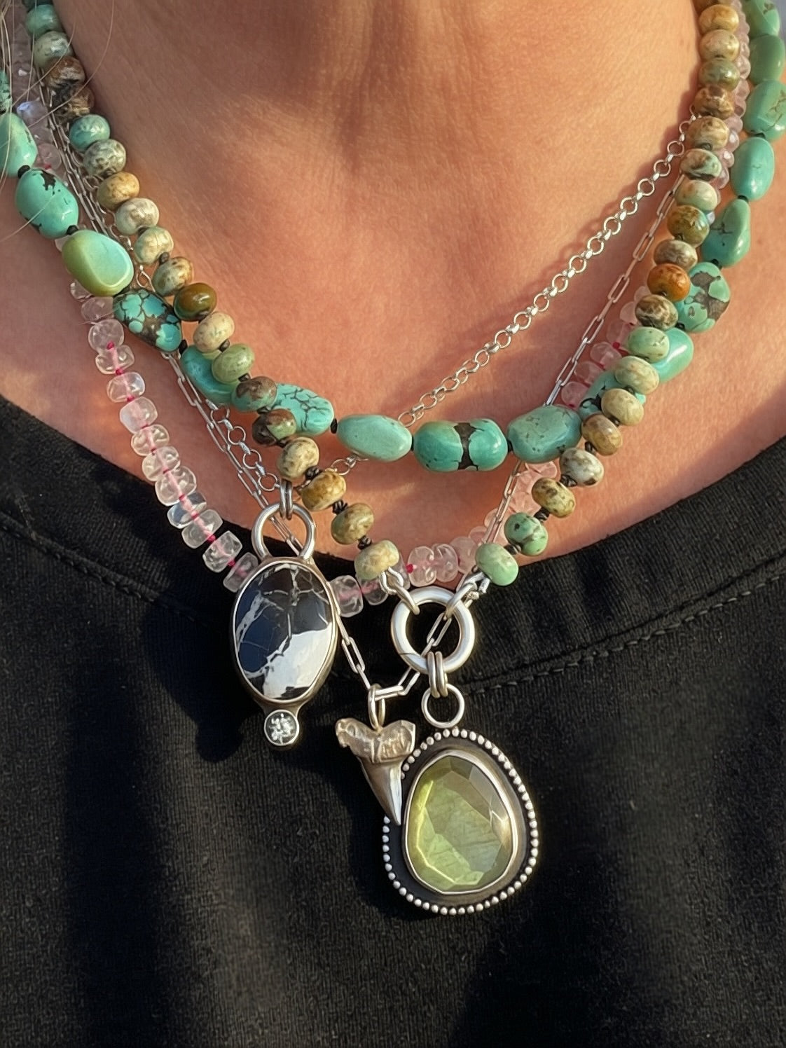 A close-up of a person wearing layered bohemian-style necklaces. The necklace features turquoise and green beads, natural stone beads, clear faceted moonstone beads on fuchsia silk, and silver charms including a shark tooth, white buffalo and spinel  and a large green labradorite pendant.
