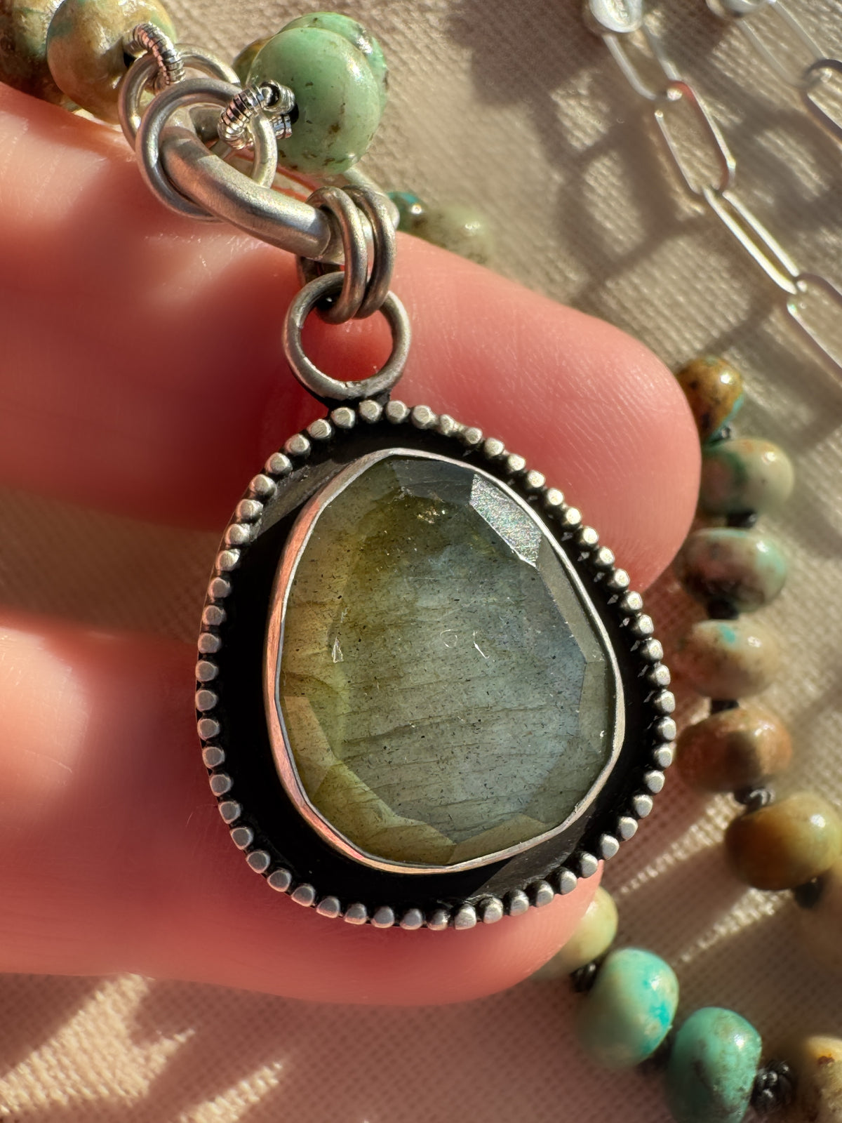 Close up of Desert Aurora necklace with turquoise beads hand-knotted on graphite silk, sterling paperclip chain and a rose-cut labradorite pendant being held.
