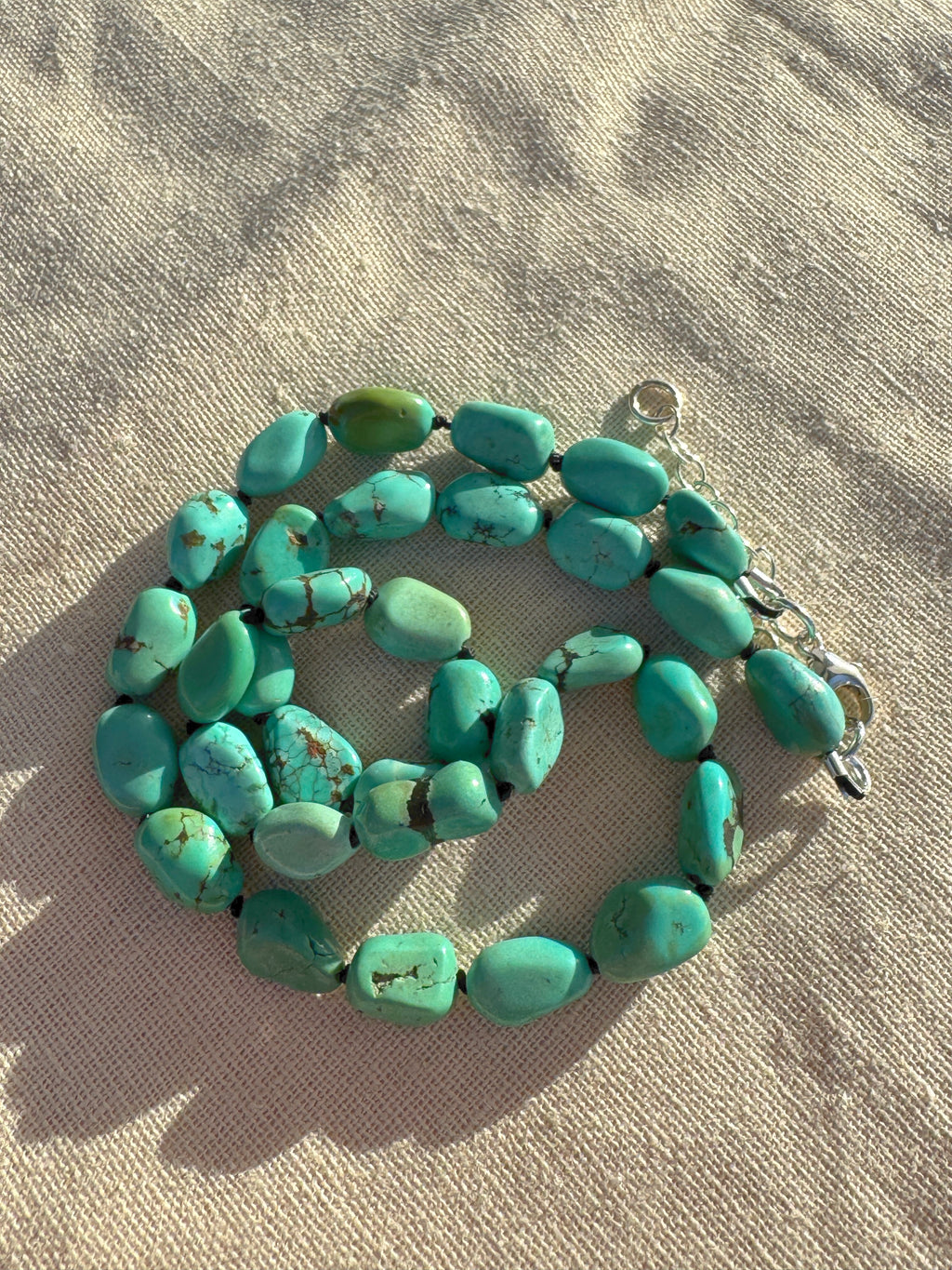 A strand of chunky turquoise beads with a silver clasp, laid out on a textured beige fabric background.