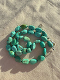 A strand of chunky turquoise beads with a silver clasp, laid out on a textured beige fabric background.