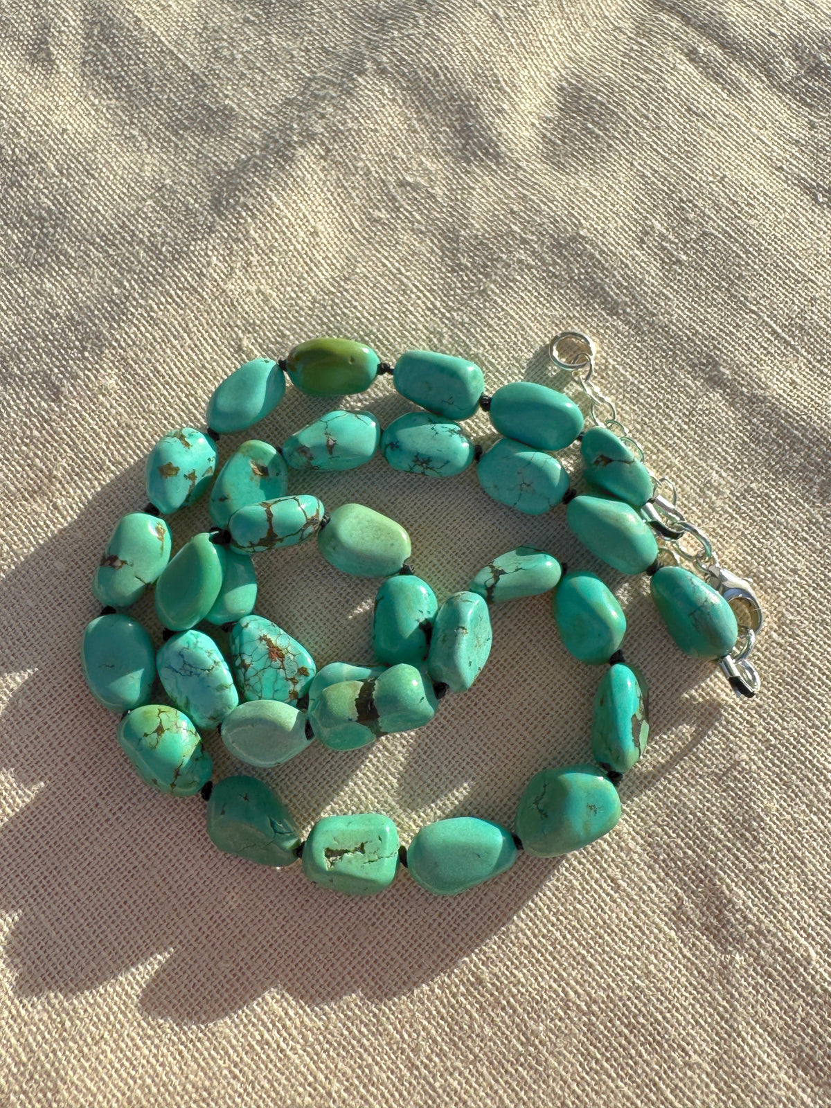 A strand of chunky turquoise beads with a silver clasp, laid out on a textured beige fabric background.