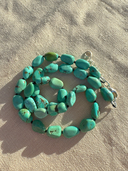 A strand of chunky turquoise beads with a silver clasp, laid out on a textured beige fabric background.