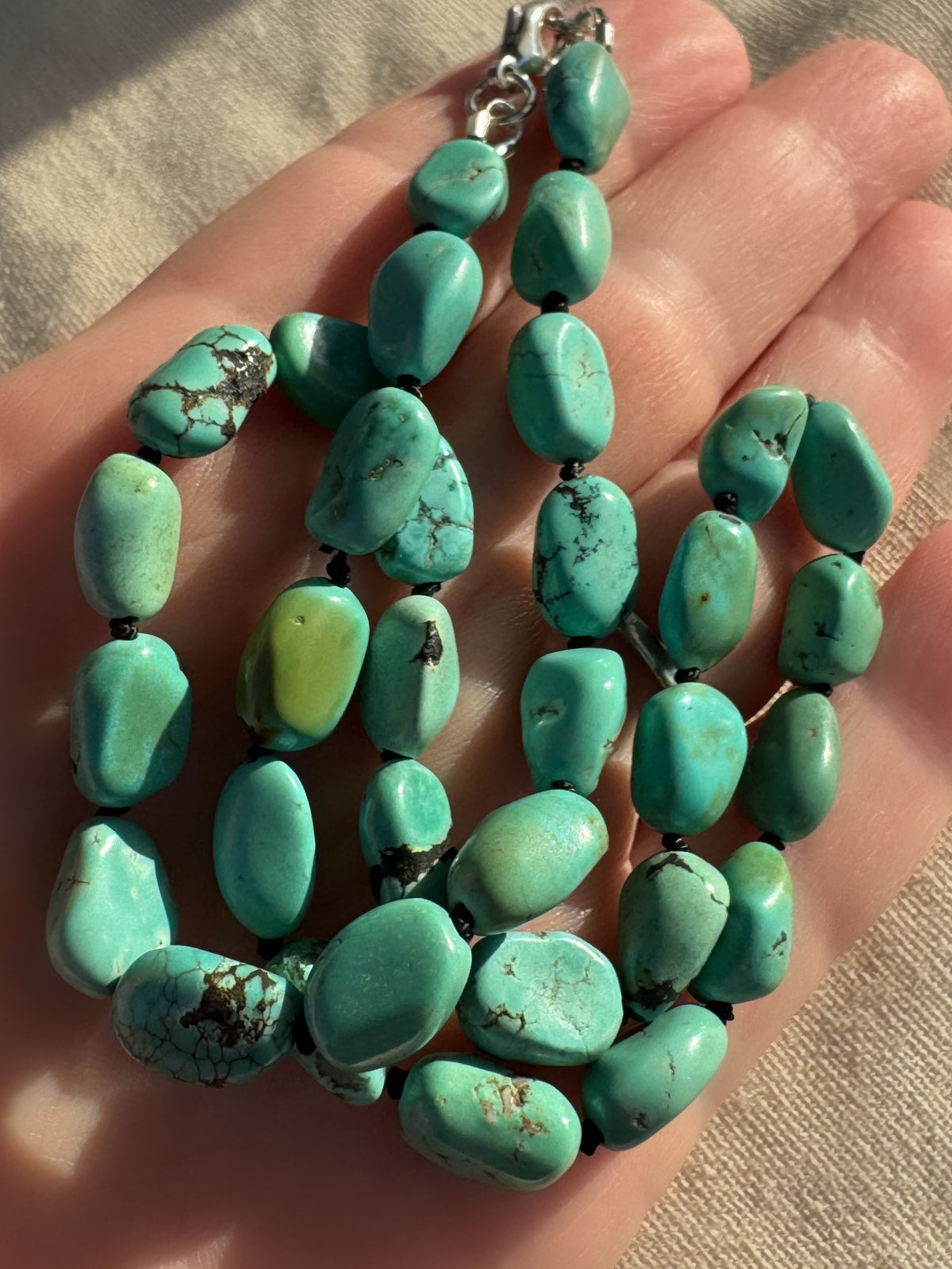 A close-up of a hand holding a strand of polished turquoise beads with a silver clasp, set against a neutral fabric background.