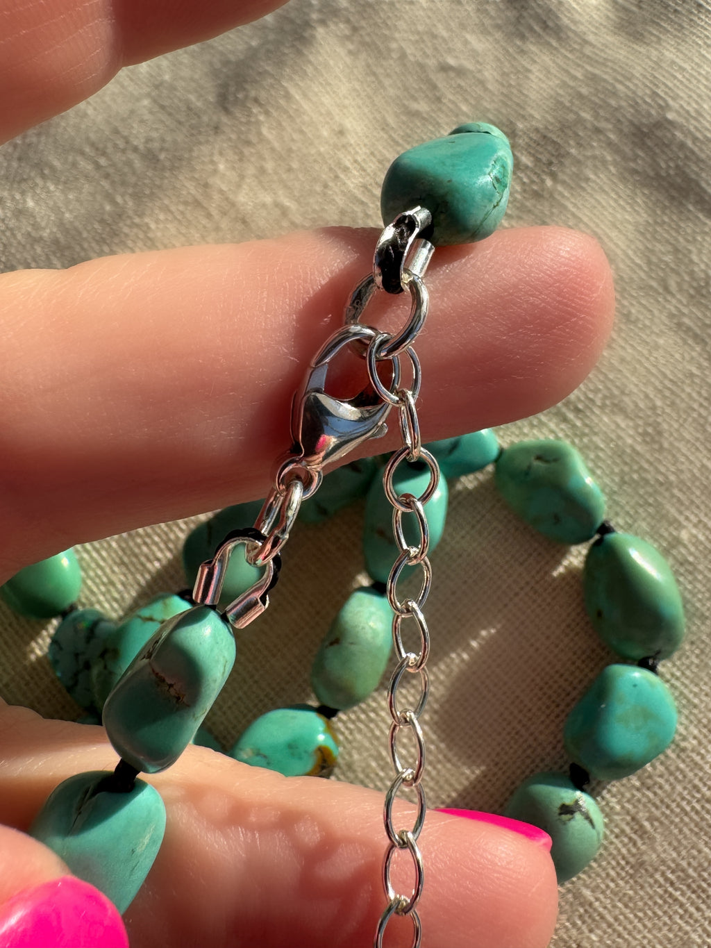 A close-up of a turquoise beaded necklace with a silver lobster clasp and extension chain, held by a hand against a neutral fabric background.