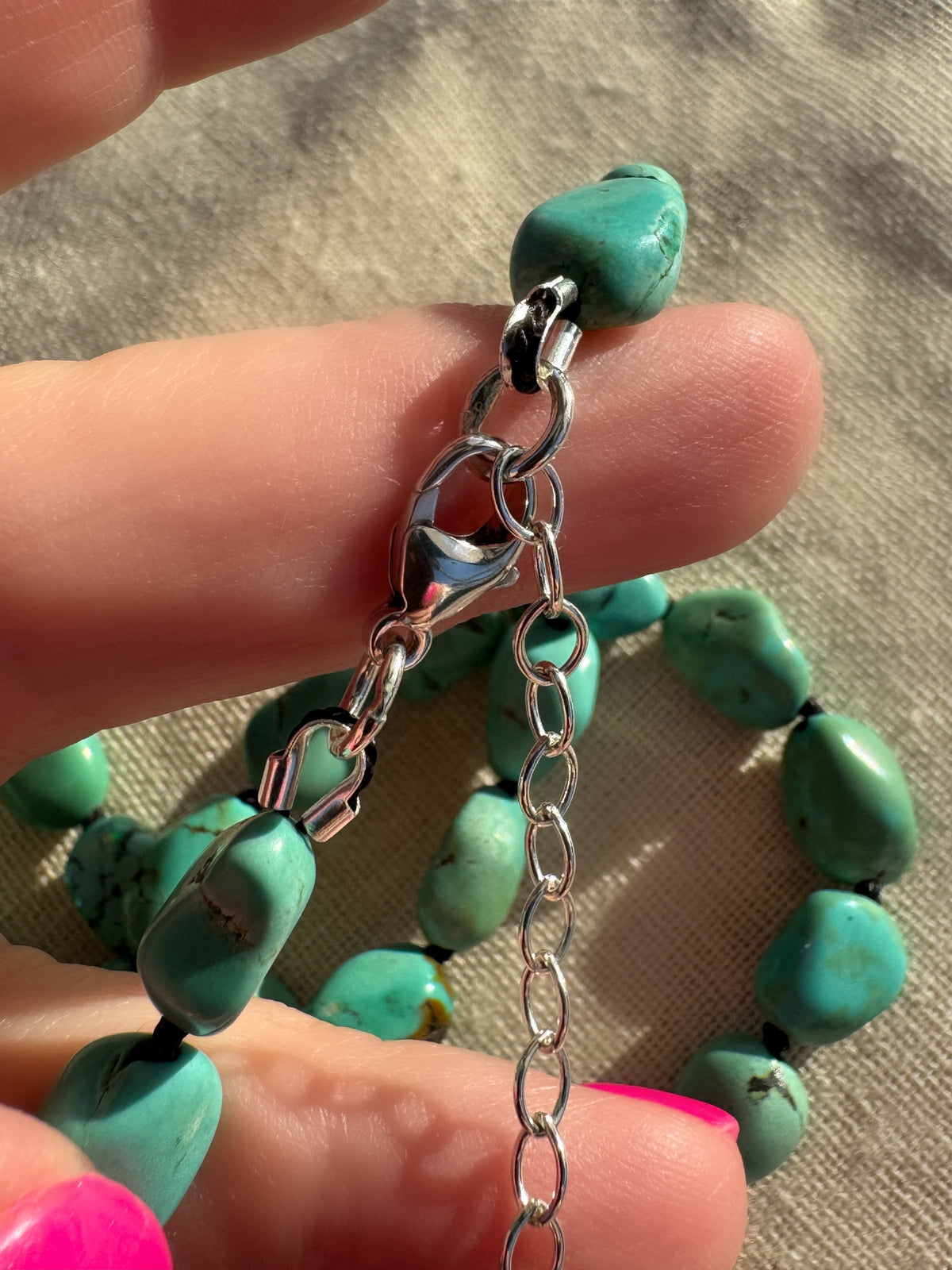 A close-up of a turquoise beaded necklace with a silver lobster clasp and extension chain, held by a hand against a neutral fabric background.