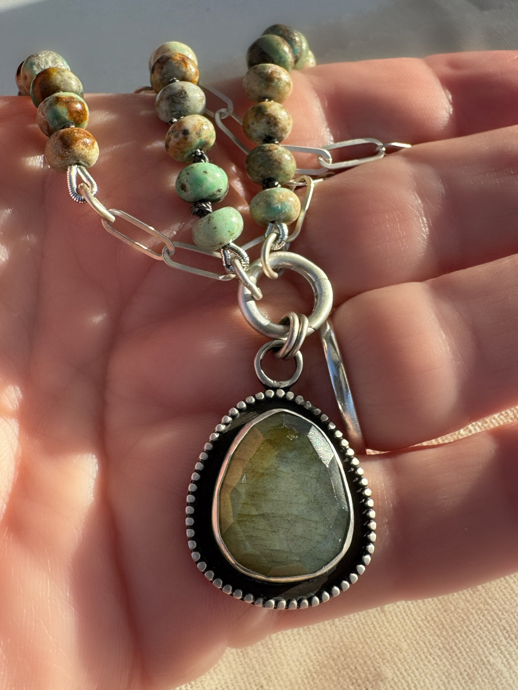 Close up of Desert Aurora necklace with turquoise beads hand-knotted on graphite silk, sterling paperclip chain and a rose-cut labradorite pendant being held.