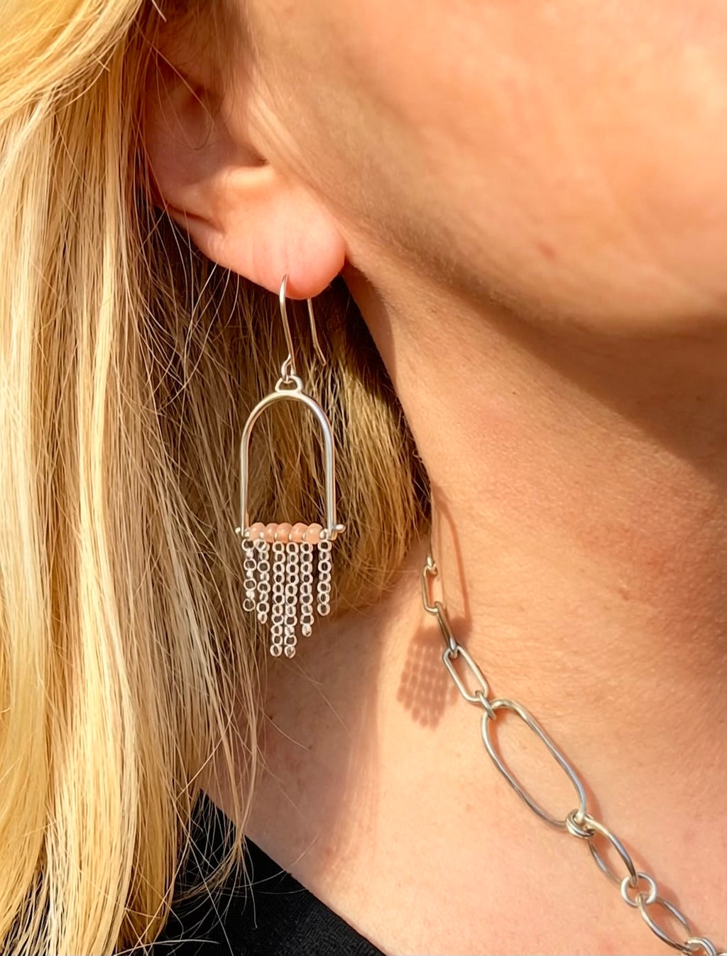 A close-up of a person's ear wearing a sterling silver drop earring with peach moonstone and sterling fringe, alongside a matching silver chain necklace.