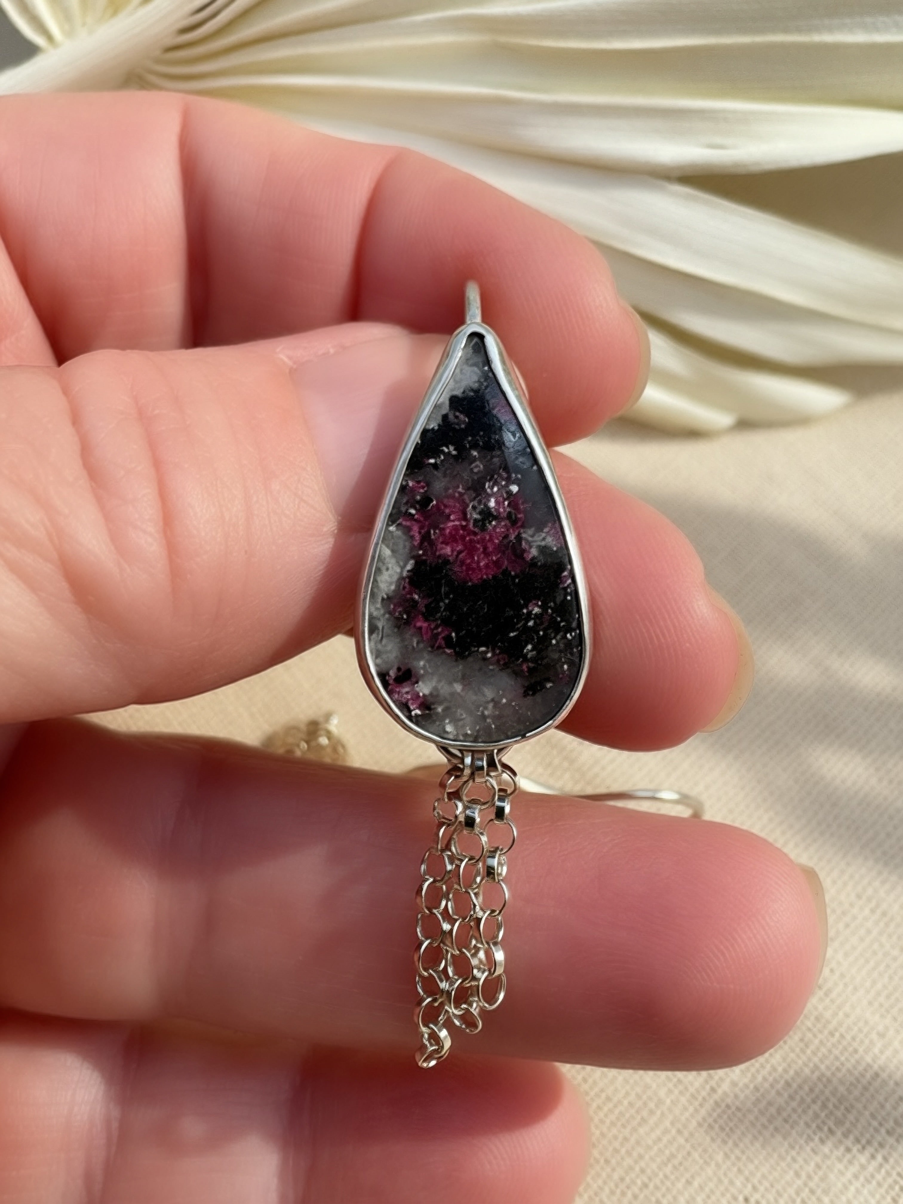 A close-up of a hand holding a teardrop-shaped silver pendant featuring a black and gray stone with pinkish-purple inclusions, suspended from a delicate silver chain.