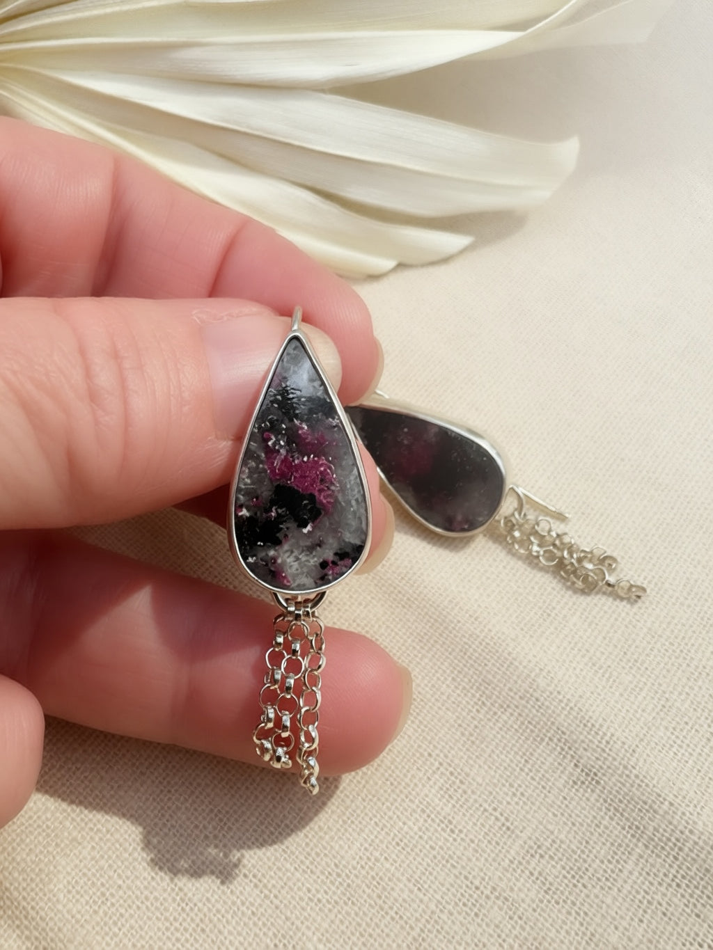 A close-up of a hand holding a pair of teardrop-shaped silver earrings with dark, cloudy gemstone centers and dangling silver chains, set against a neutral beige fabric background.