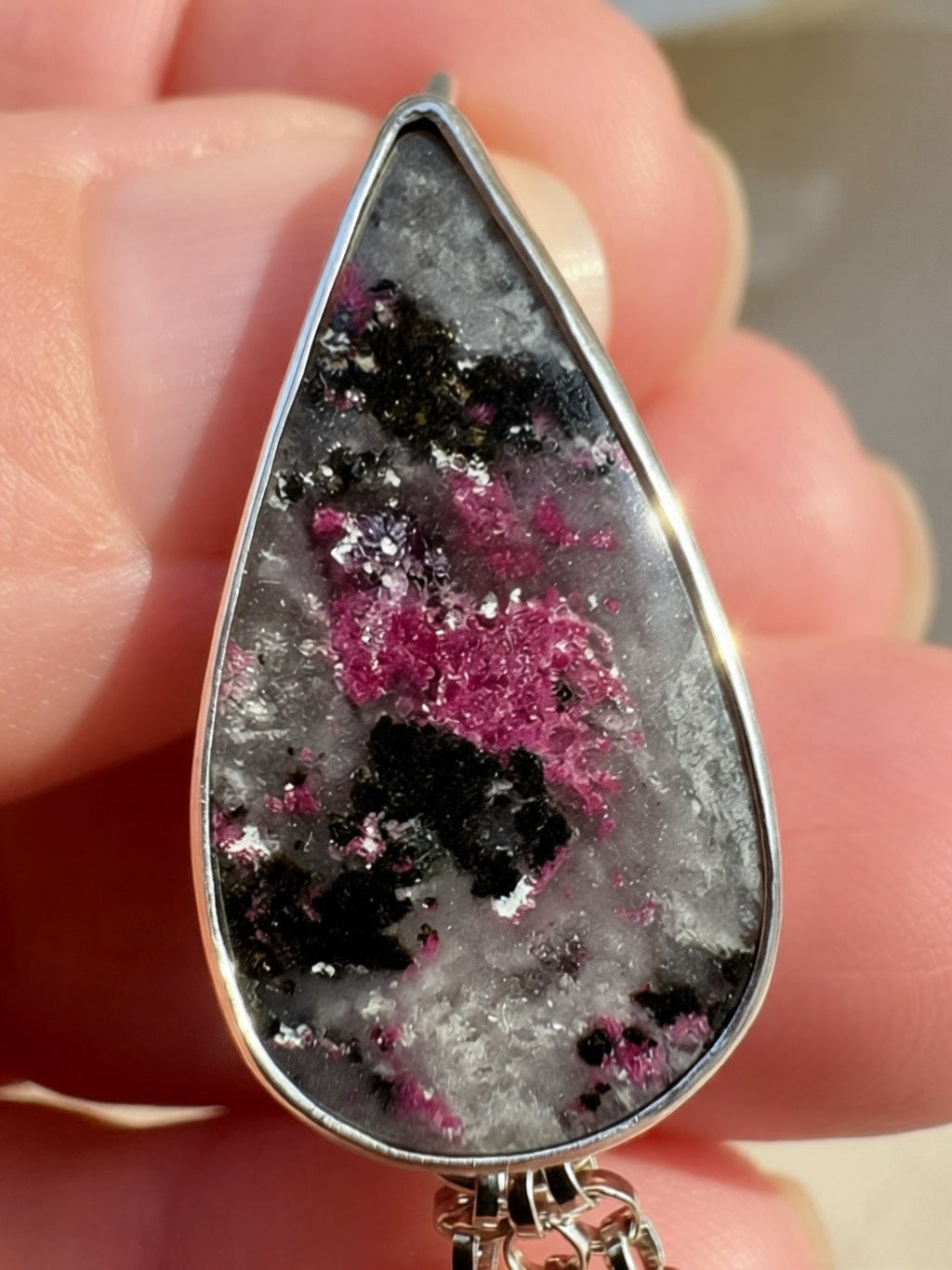 A close-up of a hand holding a teardrop-shaped pendant with a silver-toned metal border. The pendant features a natural-looking gray and white crystalline center with prominent pink and black mineral inclusions.