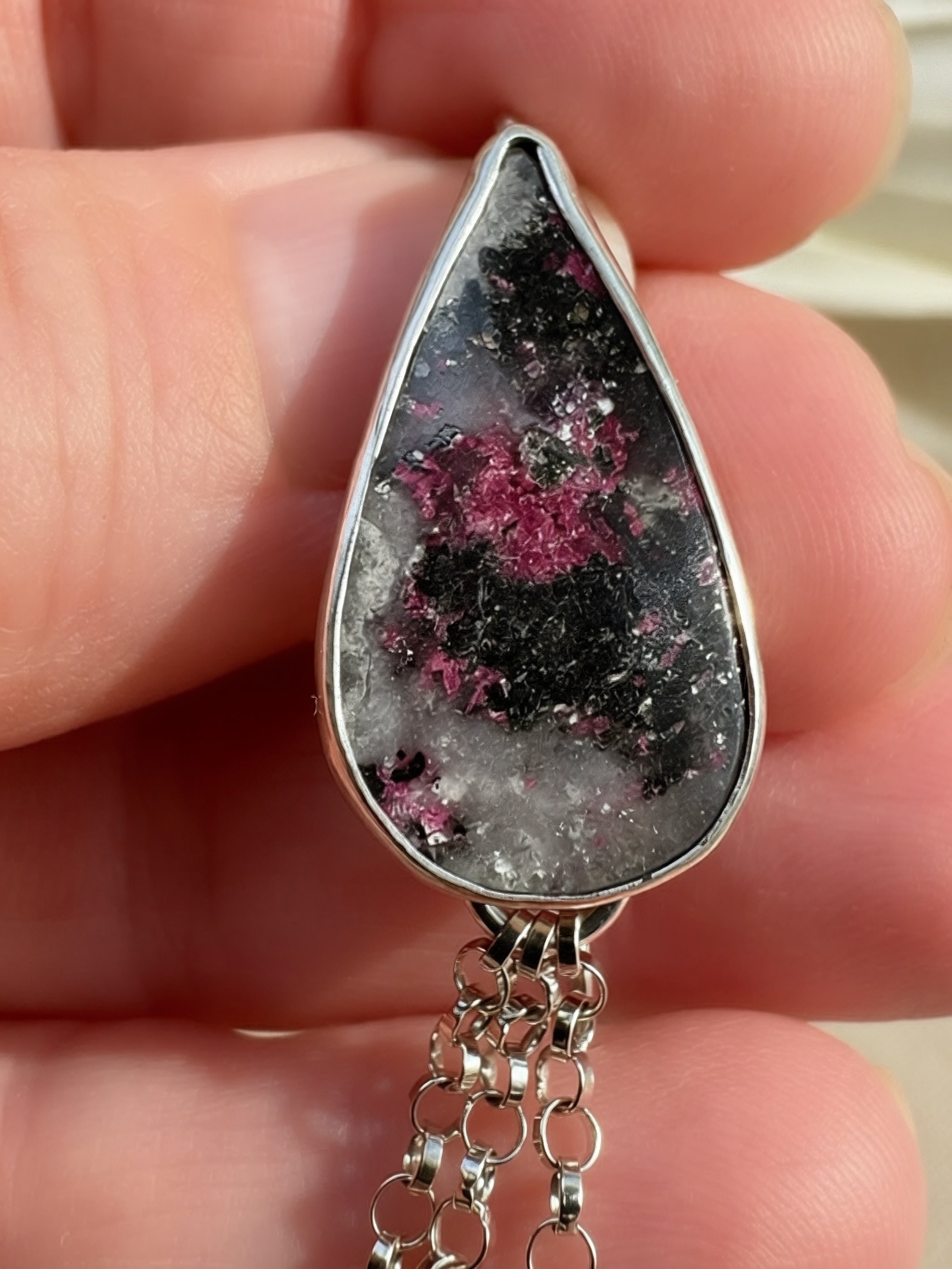 A close-up of a hand holding a teardrop-shaped silver pendant featuring a black and white geode-like pattern with vibrant pink mineral inclusions, with silver fringe
