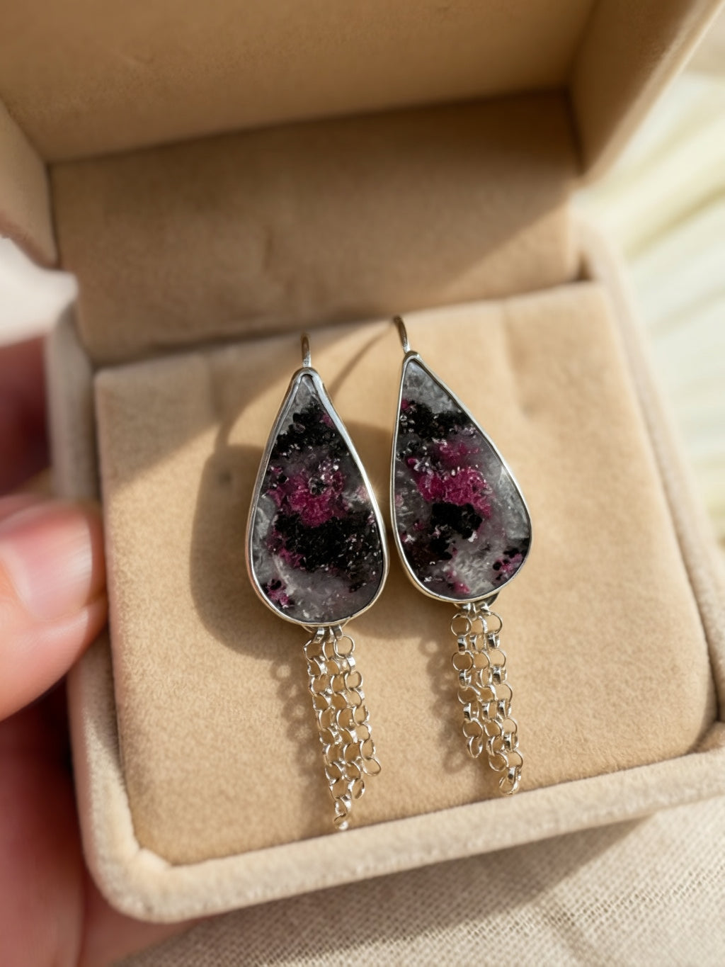 A pair of teardrop-shaped dangle earrings with stones with black, white and hot pink inclusions, with silver fringe chains inside a beige jewelry box. A hand is holding the box on the left.