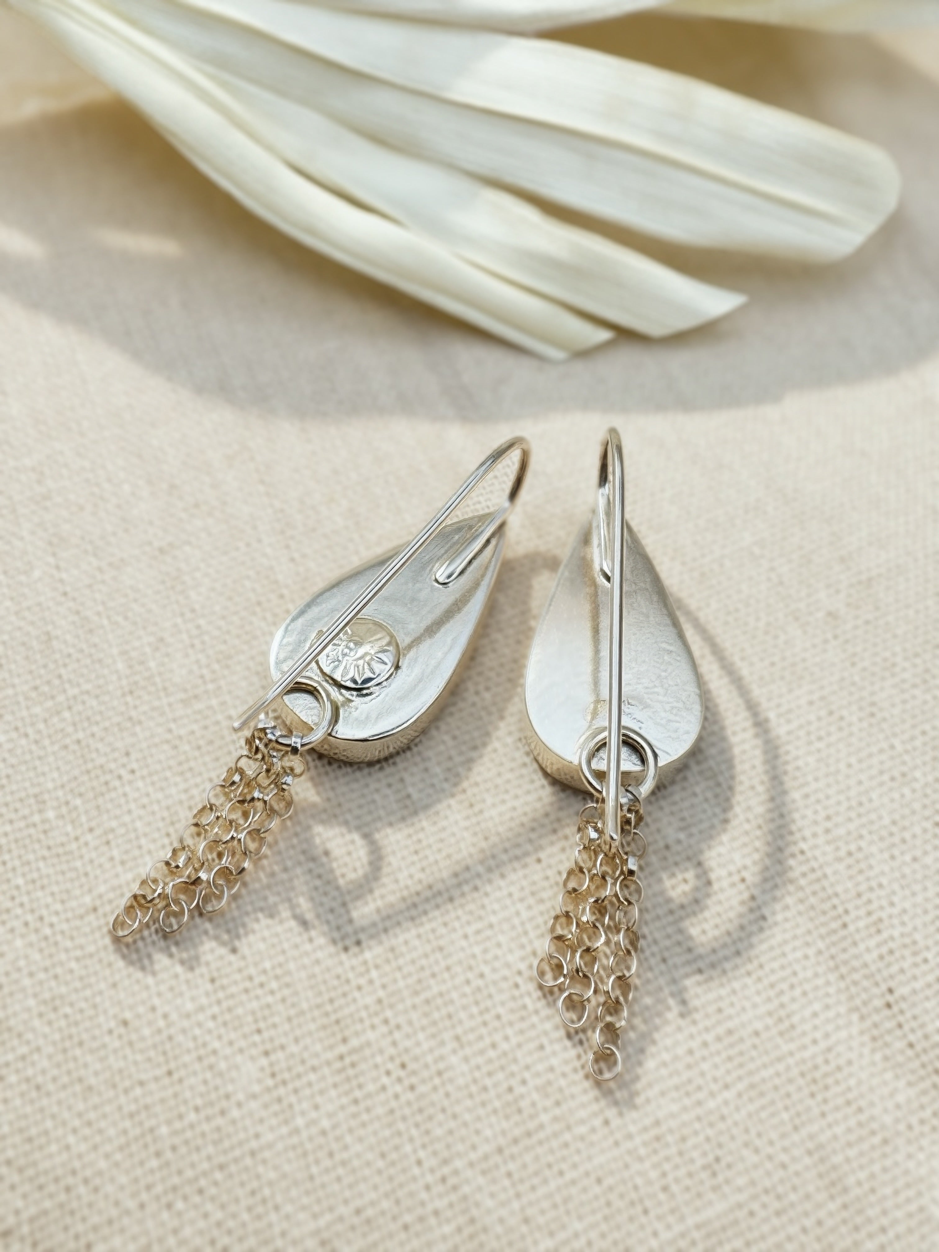 A back view of a pair of silver teardrop-shaped dangle earrings. Displayed on a textured beige fabric background with a white dried leaf in the upper corner.