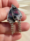 A pair of teardrop-shaped dangle earrings with silver settings and dangling chain drops, featuring a black, gray, and pink marbled stone design, held in a person's hand against a light beige fabric background.