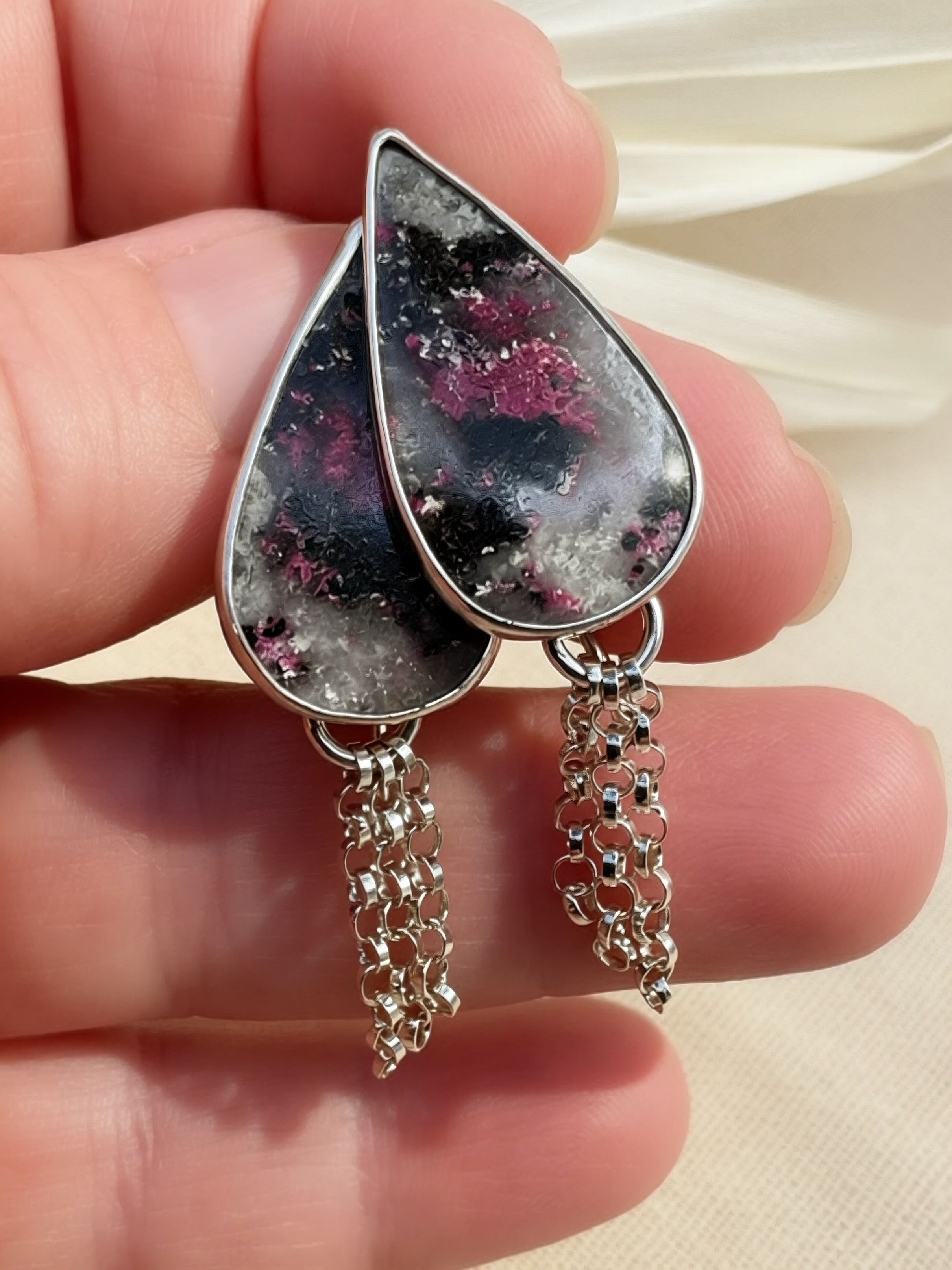 A pair of teardrop-shaped dangle earrings with silver settings and dangling chain drops, featuring a black, gray, and pink marbled stone design, held in a person's hand against a light beige fabric background.