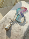 A beaded necklace with a baroque pearl pendant, featuring translucent beads in pastel rainbow colors and a sterling silver chain, displayed on a textured beige fabric.
