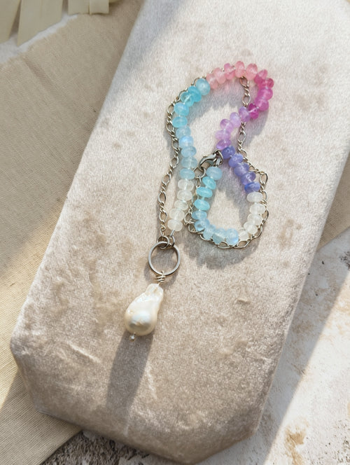 A delicate beaded necklace with pastel blue, pink, and purple beads, a silver chain, and a white baroque pearl pendant, displayed on a textured beige fabric surface.