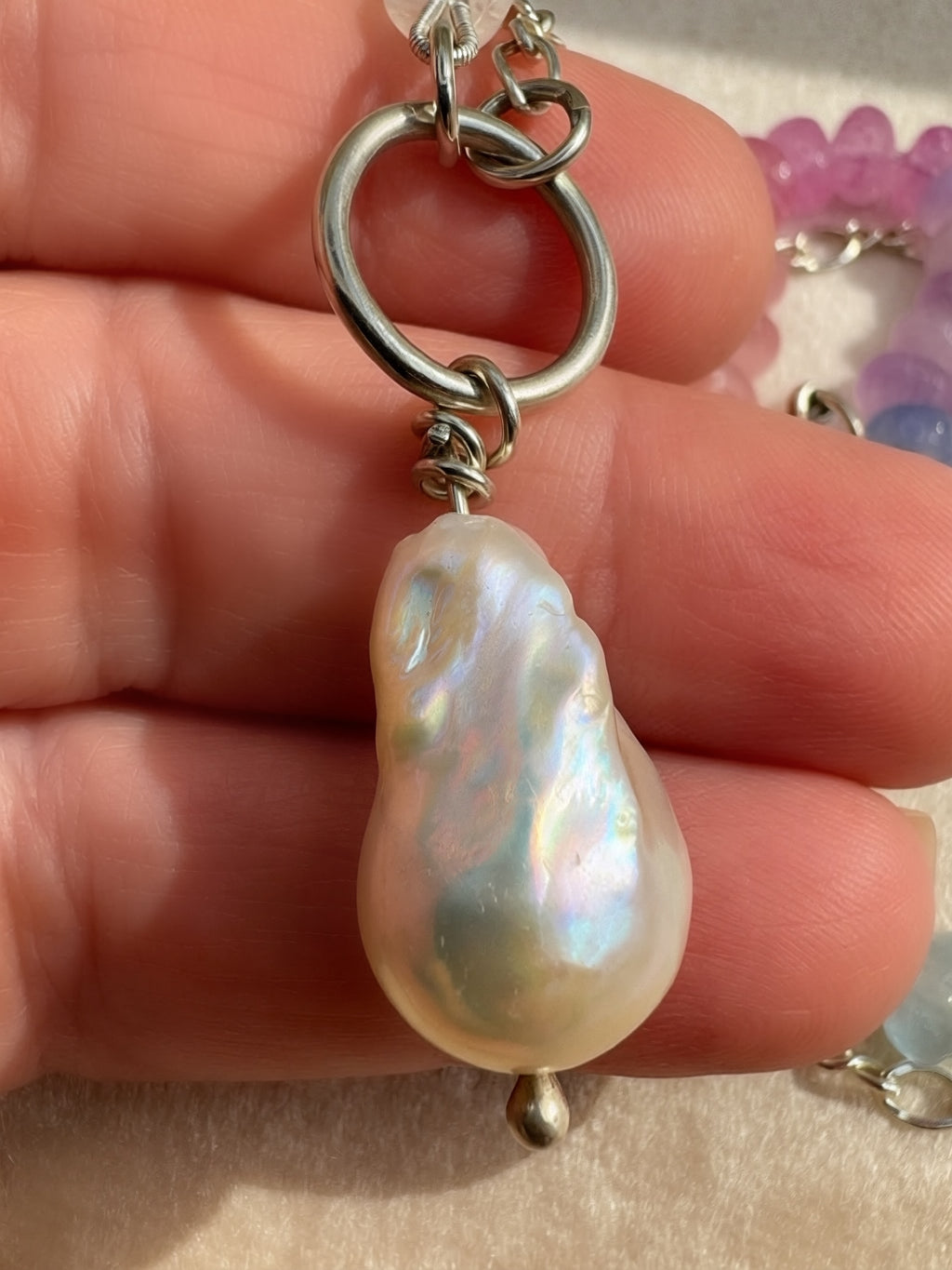 A close-up of a hand holding a silver and beaded necklace featuring a large, irregular baroque pearl with an iridescent white sheen.