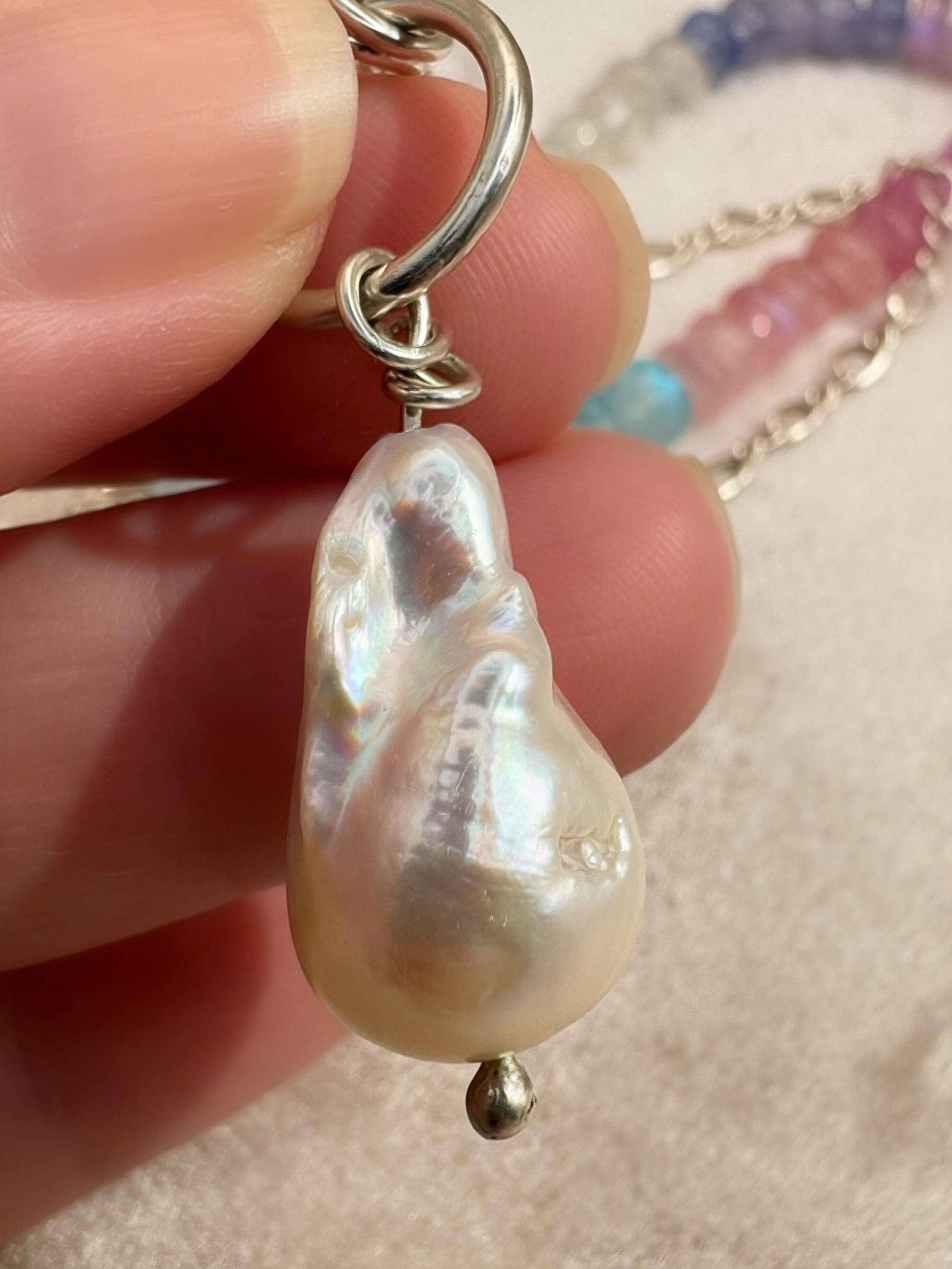 A close-up of a hand holding a baroque pearl pendant necklace with a silver clasp and chain, with the beaded necklace blurred in the background.