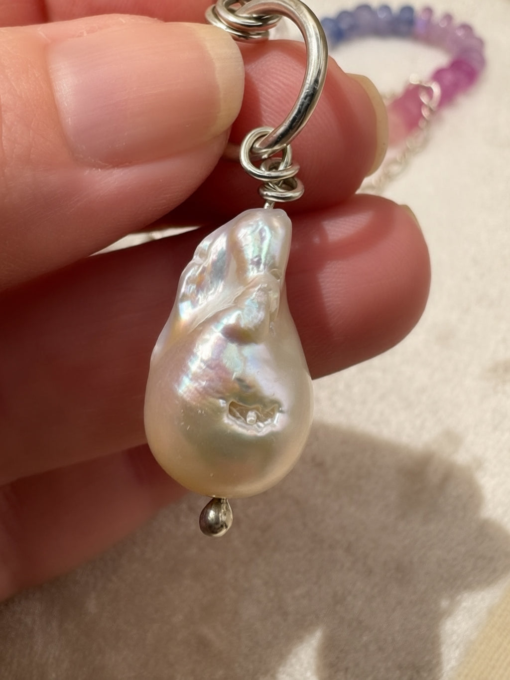 A close-up of a hand holding a baroque pearl pendant with a silver clasp, with the beaded necklace visible in the background.