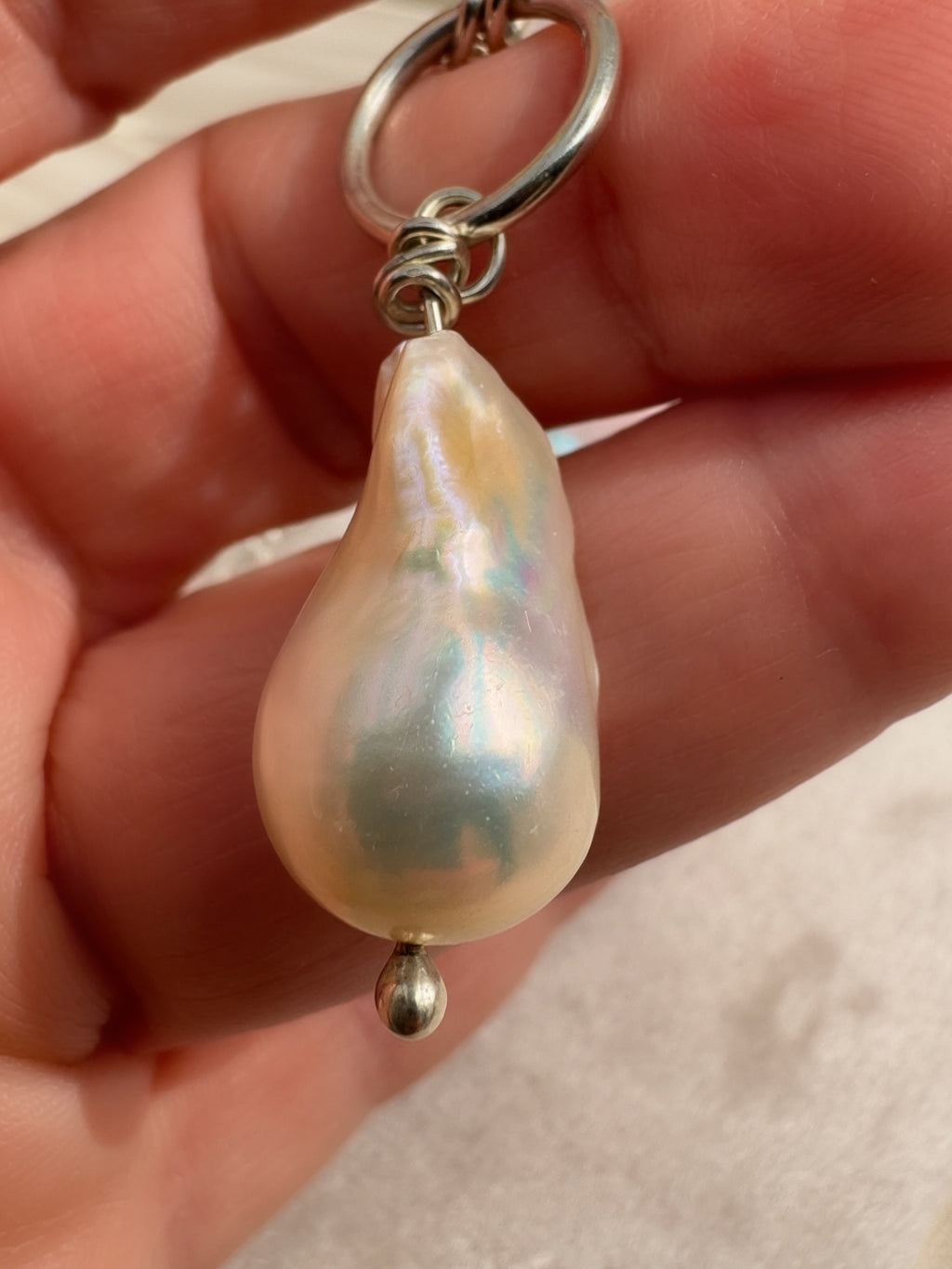 A close-up of a hand holding a silver chain with a large, irregular baroque pearl pendant that has iridescent pearlescent colors.